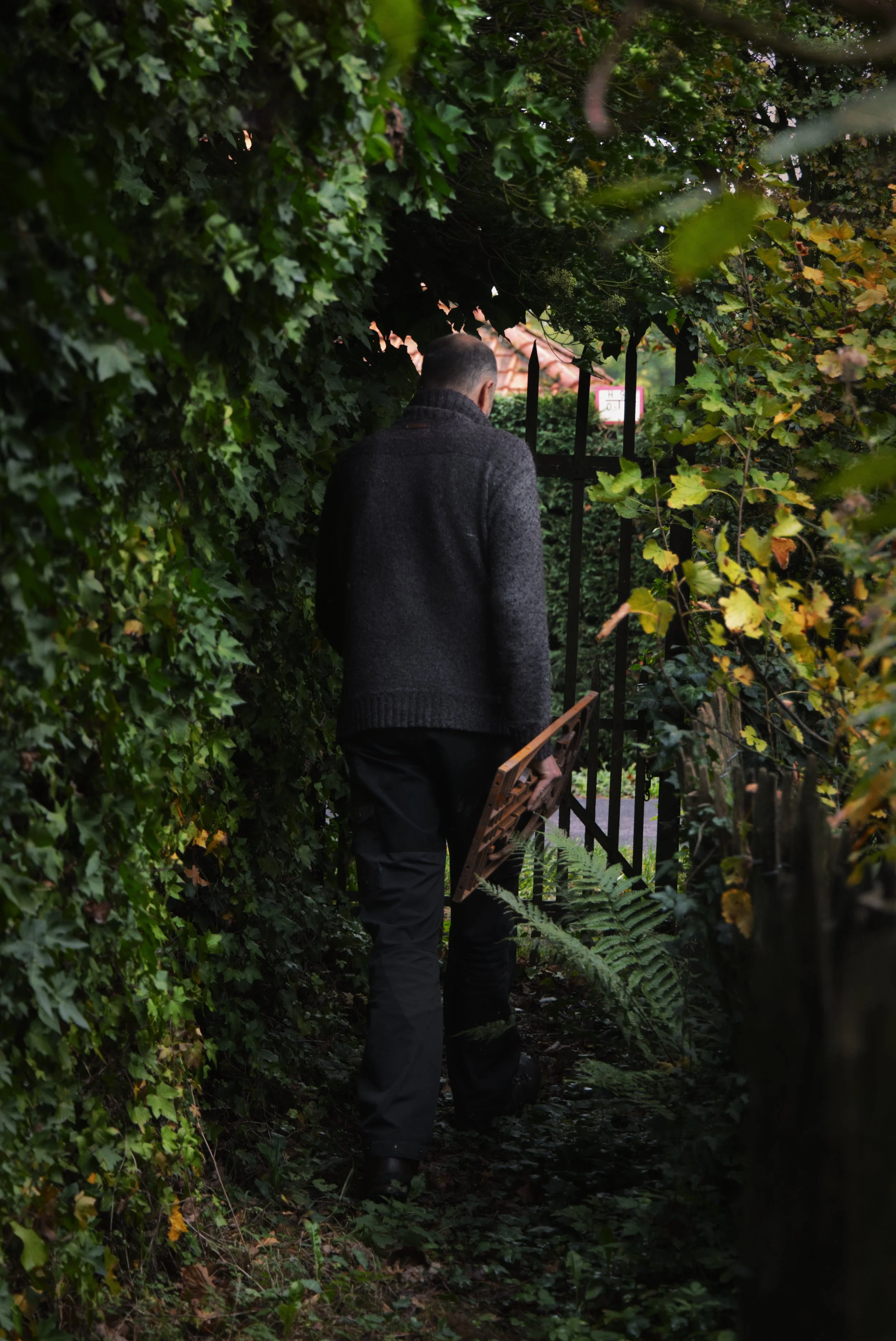 Man met een jas en broek, die een stuk tuinhek of hekwerk draagt, loopt door een smal pad tussen dichtbegroeide struiken en bomen. Hij houdt een weefstoeltje in zijn hand.