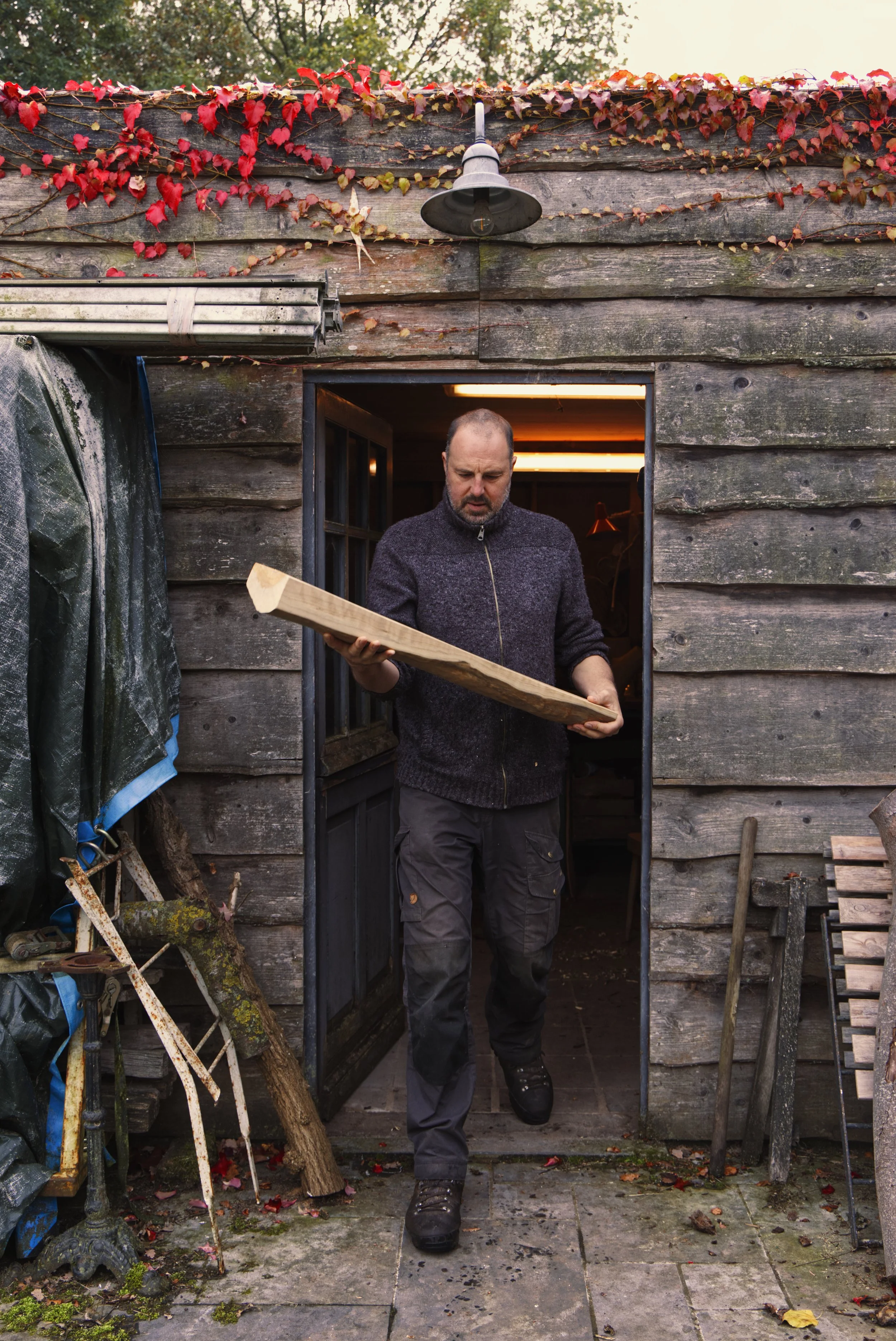 Man die een stuk hout vasthoudt voor de deur van een houten schuur, buiten, omgeven door herfstbladeren en tuingereedschap.
