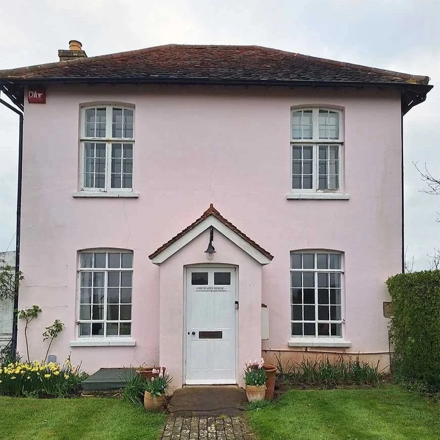 A pink two-story house with four large white-framed windows, a white door with a letter slot, a small gable roof above the door, and a garden with potted plants in front.