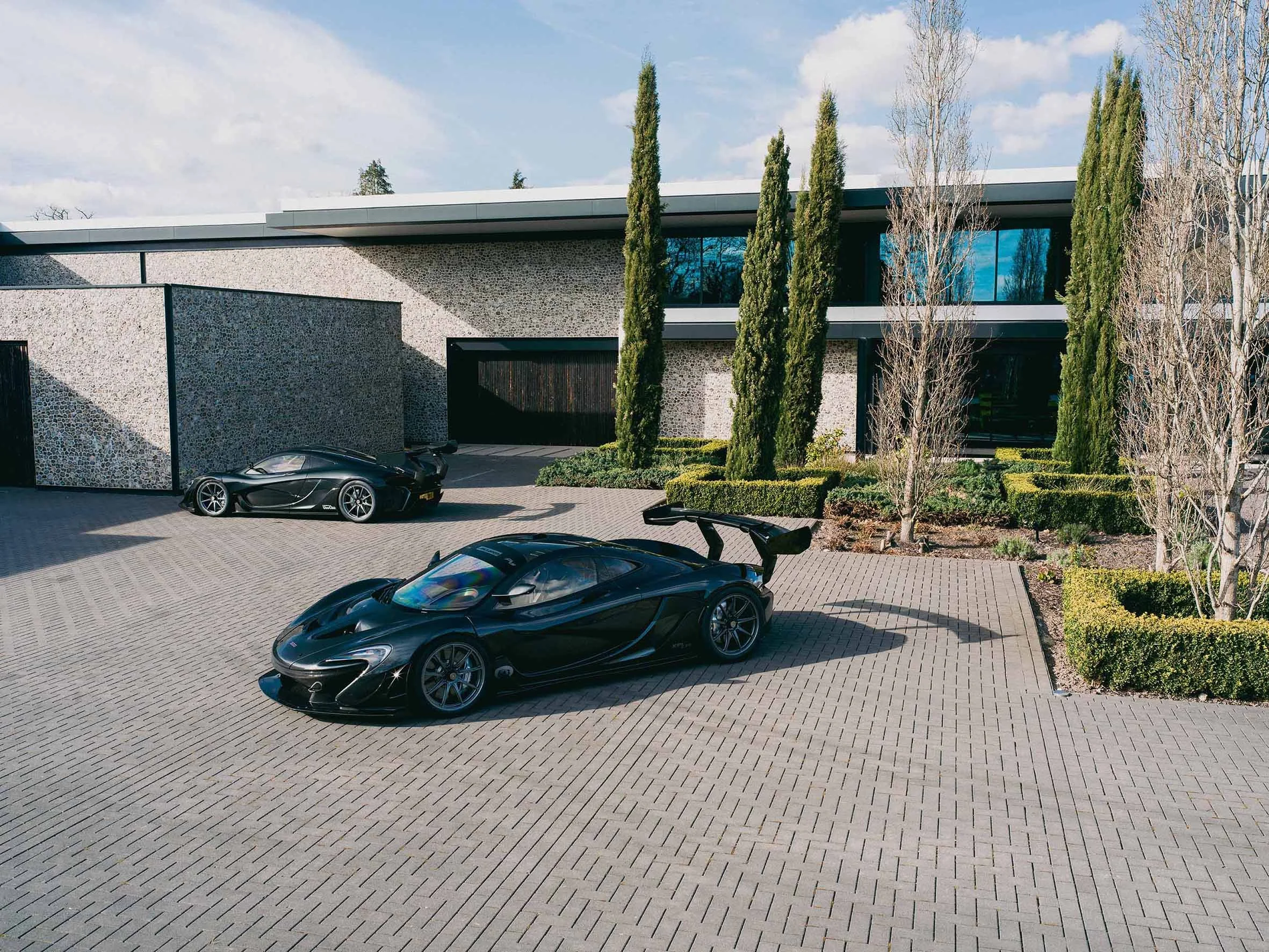 Luxury black sports cars parked in a driveway in front of a modern house with large windows and tall trees, under a partly cloudy sky.