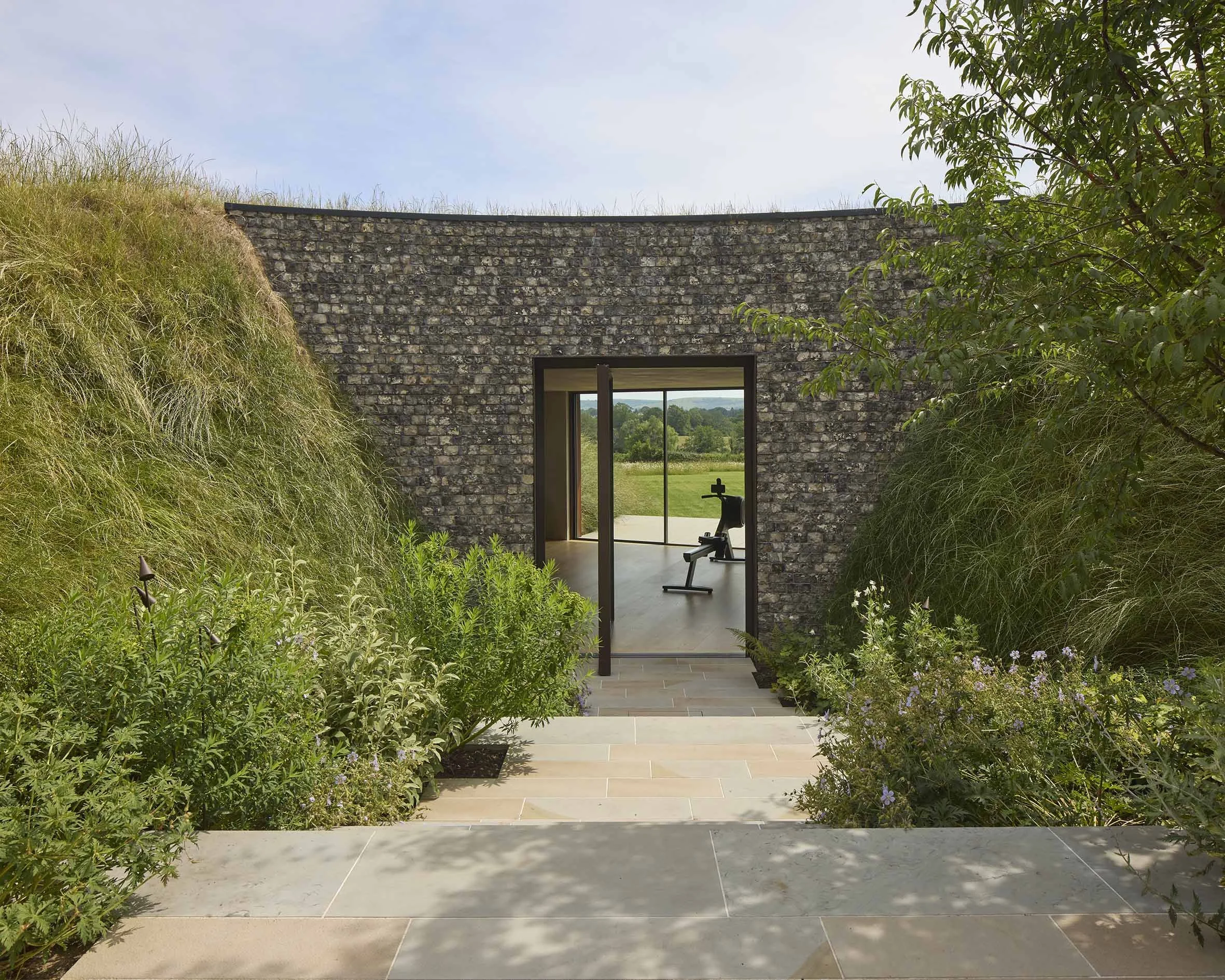 A modern stone house with glass doors opening to a lush green garden and a view of fields beyond. The area is framed by tall grasses and various plants, with a clear blue sky overhead.