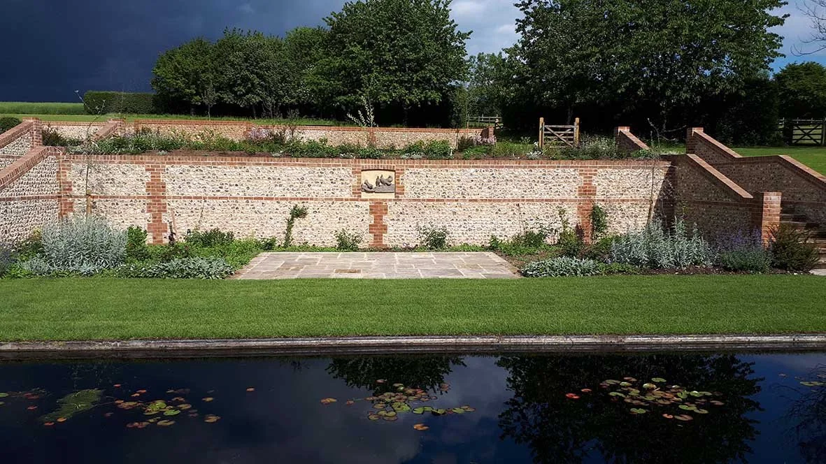 A landscaped garden with stone and brick walls, stairs, and greenery, beside a pond with water lilies.