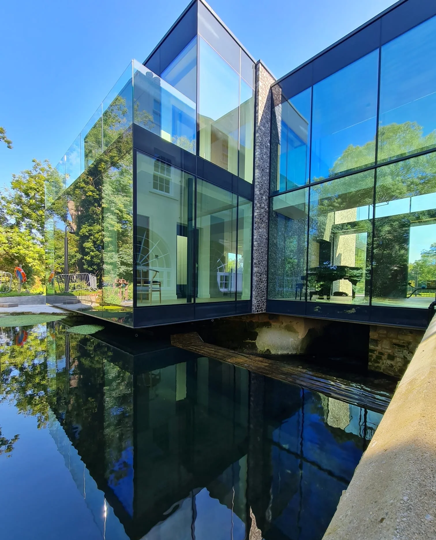 A modern building with glass walls reflecting trees and the blue sky, situated over a pond with a floating reflection.