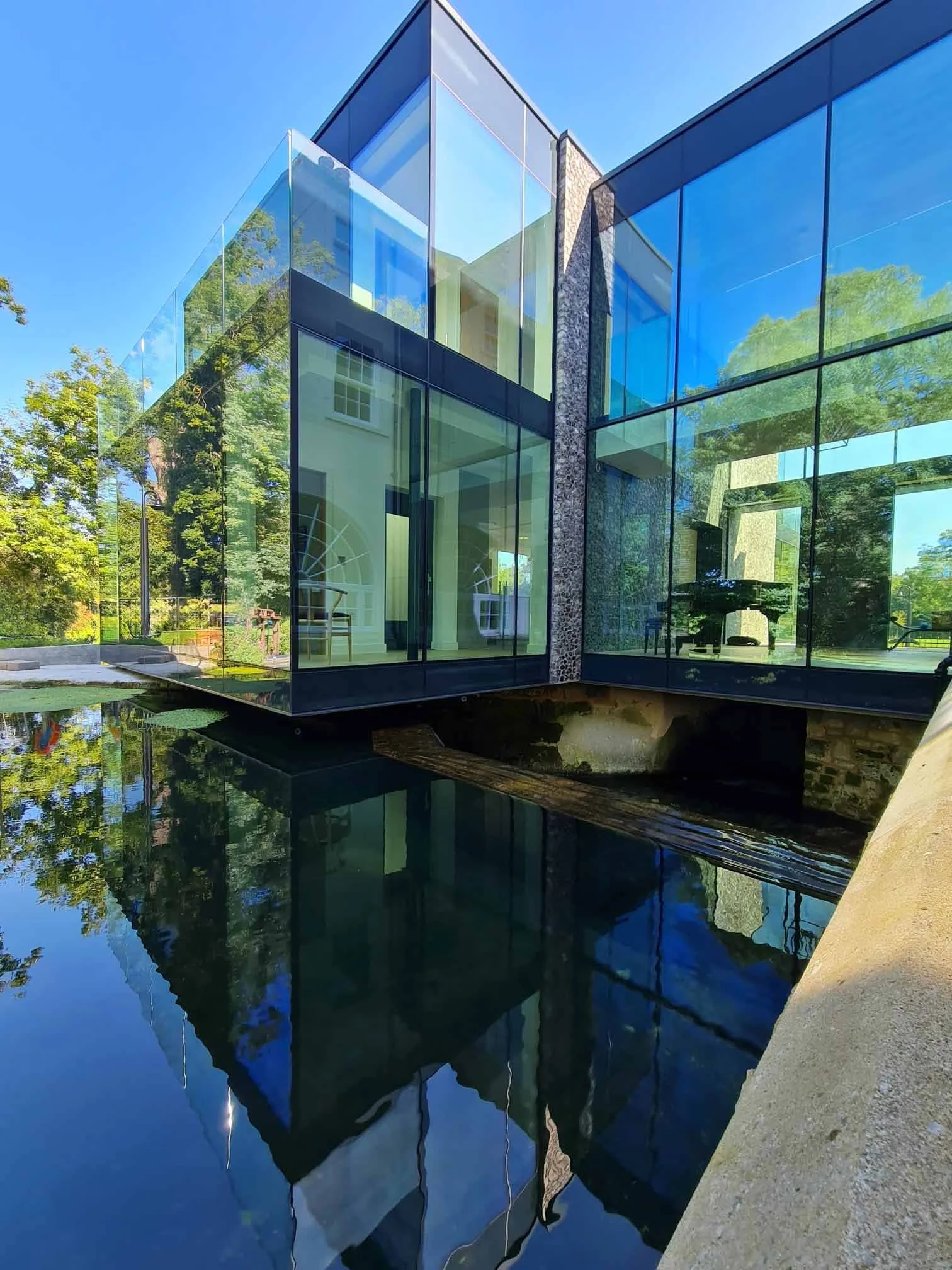 flint and glazed extension with blue sky and trees reflected surrounded by water