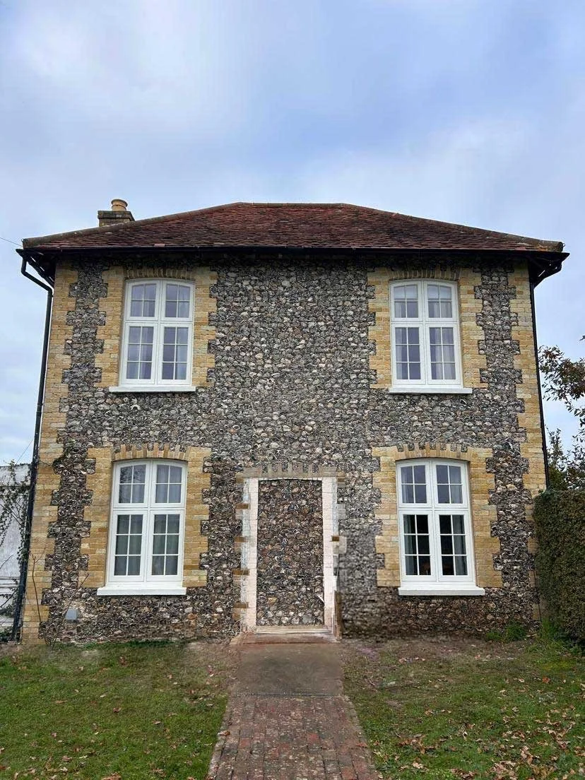A two-story house with a stone exterior, four white-framed windows, and a red-tiled roof. The front entrance is boarded up with wooden planks.