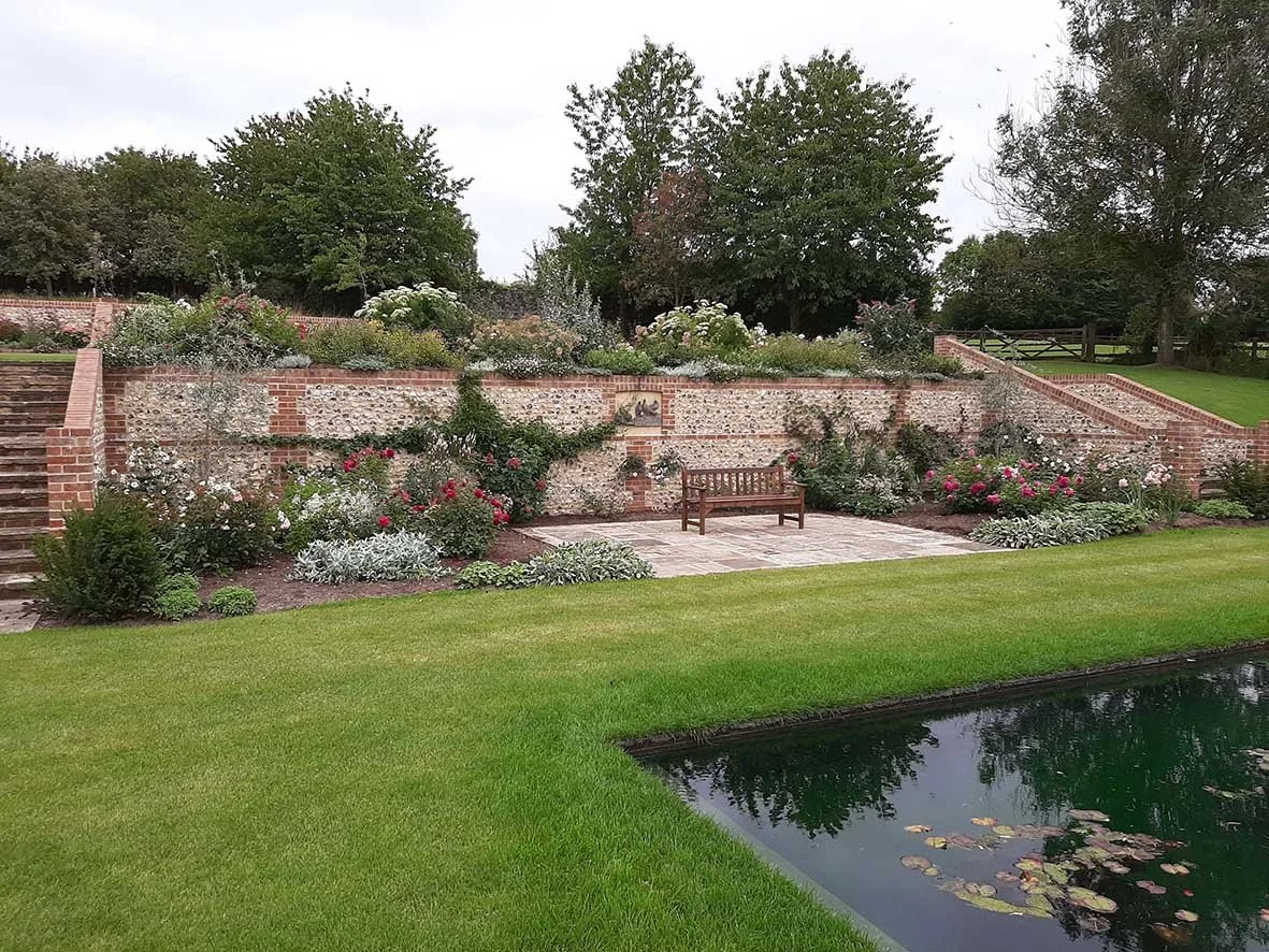 A landscaped garden with a small pond, a grassy lawn, and a brick terraced retaining wall filled with flowering plants and bushes, with some trees in the background.