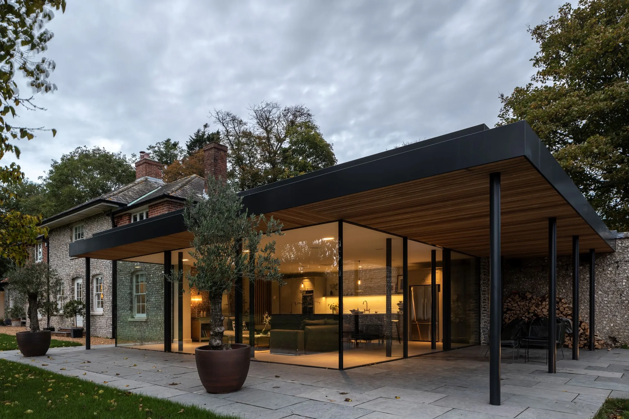 Modern house with glass walls and wooden ceiling extension, surrounded by potted trees, with autumn-colored trees and a cloudy sky in the background.