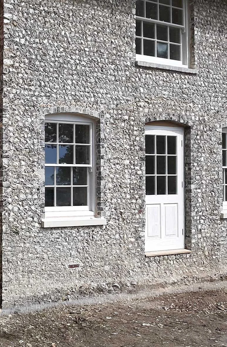 Close-up of a stone house wall with three white-framed windows and a white door, all with multiple panes. The door is below a window, and the ground in front is unpaved.