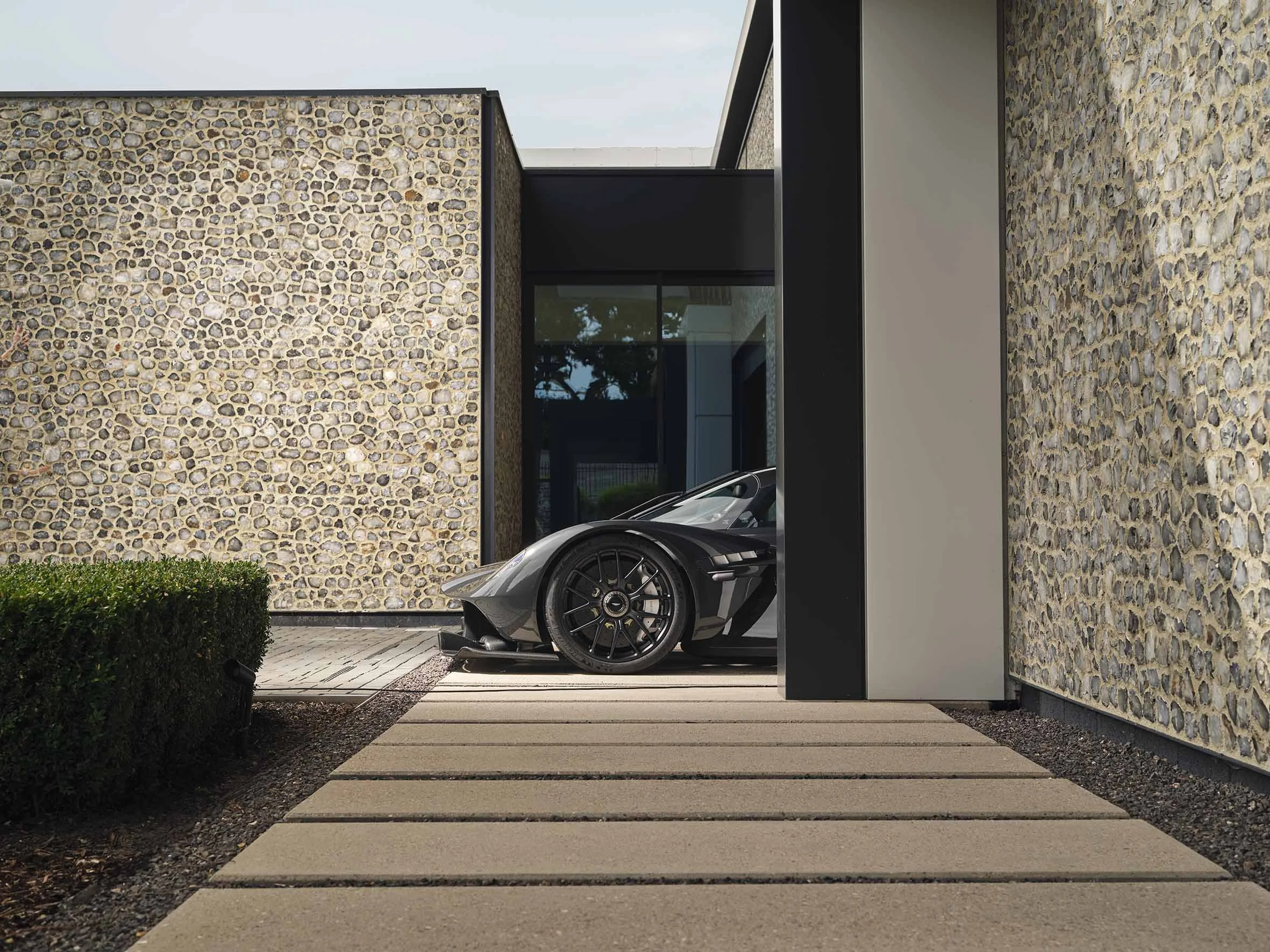 View of a modern house with stone and smooth wall finishes, seen through a driveway with concrete slabs, and a luxury sports car parked underneath a carport.