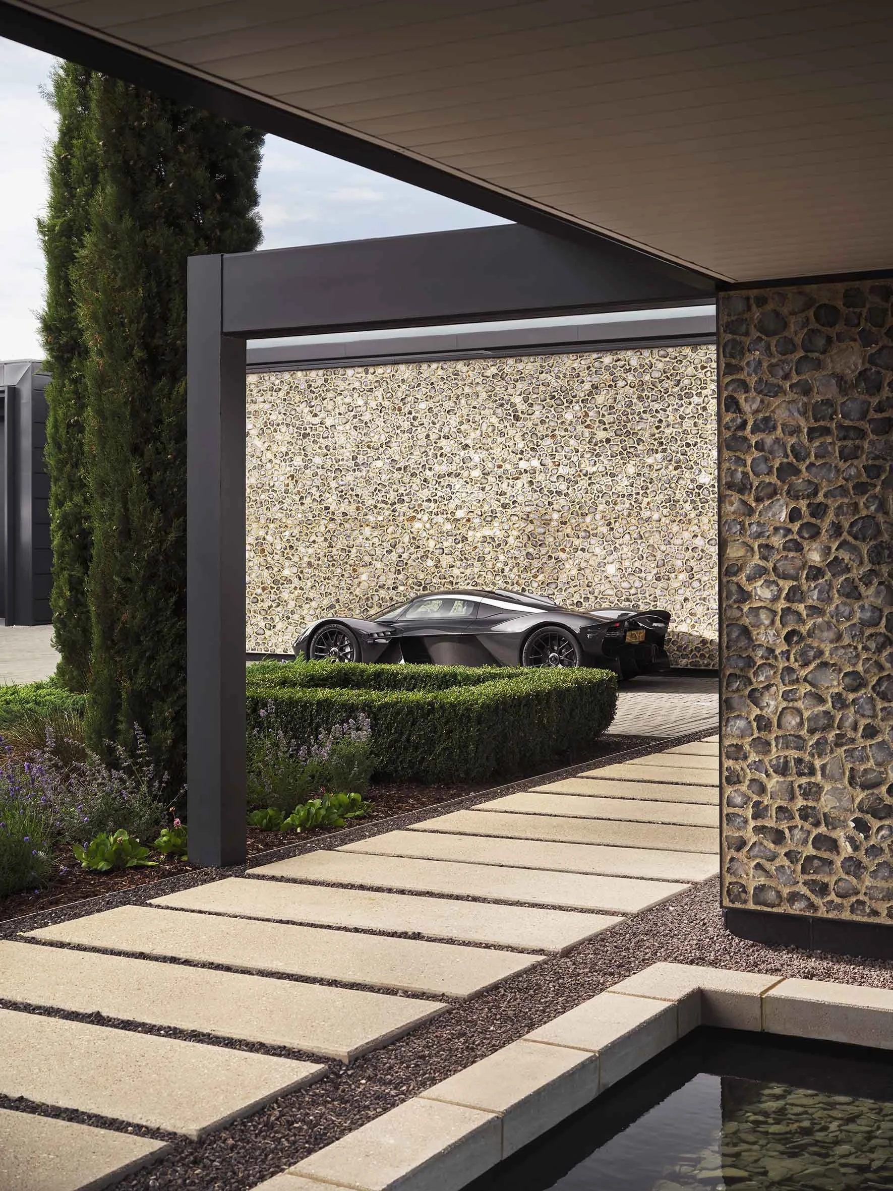 Modern outdoor area featuring a black sports car parked against a pebble stone wall, with a landscaped garden in the foreground and sleek architectural elements.