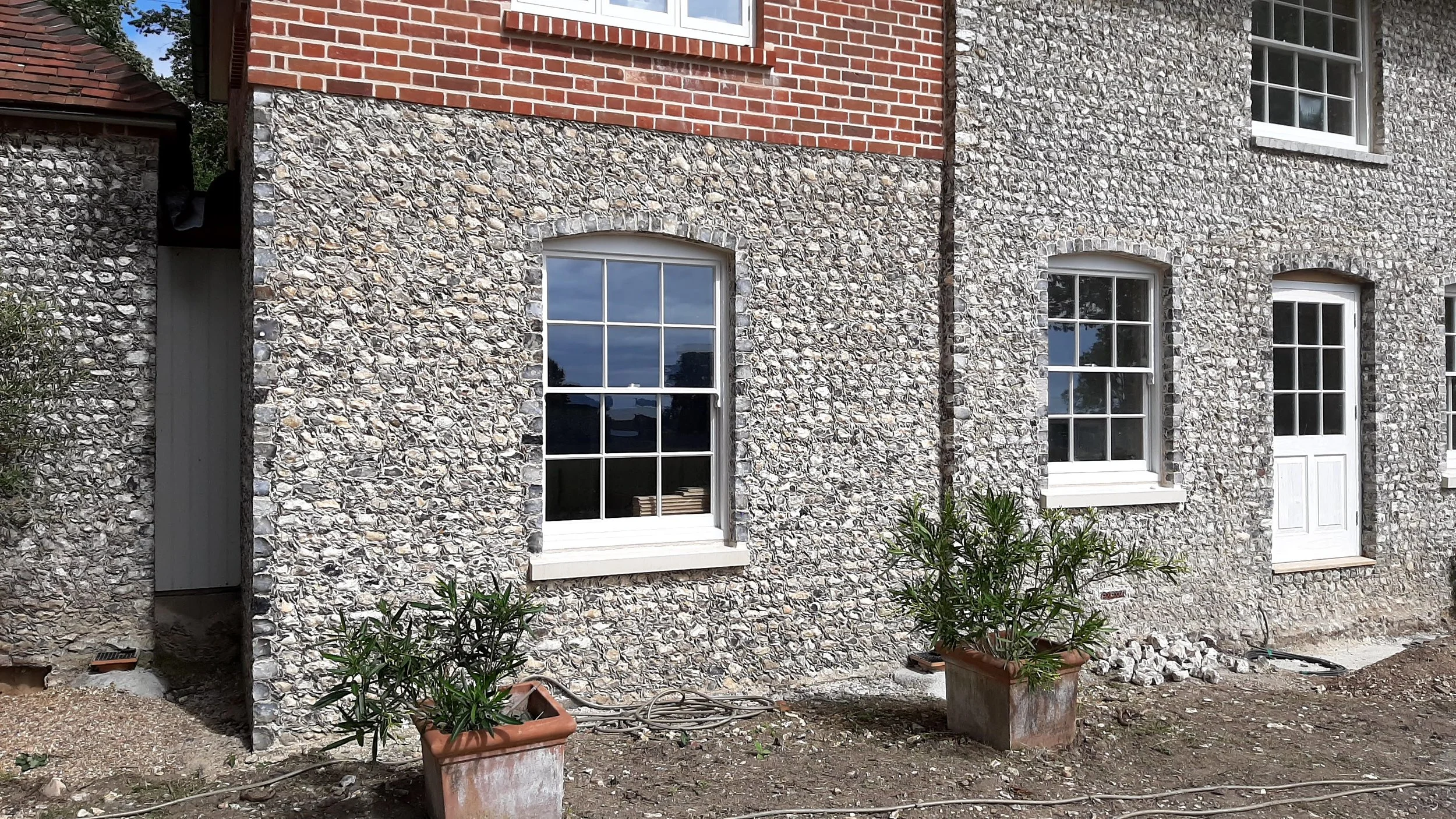 Exterior view of a building with a mixed brick and cobblestone facade, three large windows with white frames, and a white door. There are potted plants with greenery and some gardening supplies on the ground outside.