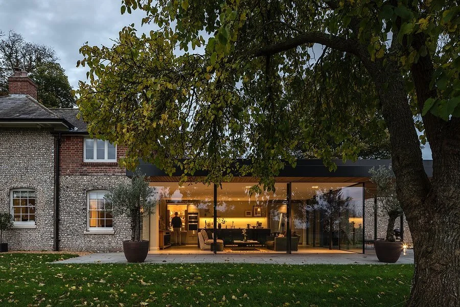 Modern glass extension attached to an older stone house, illuminated from inside, with large potted plants and a grass lawn with fallen leaves in the foreground, and mature trees around.