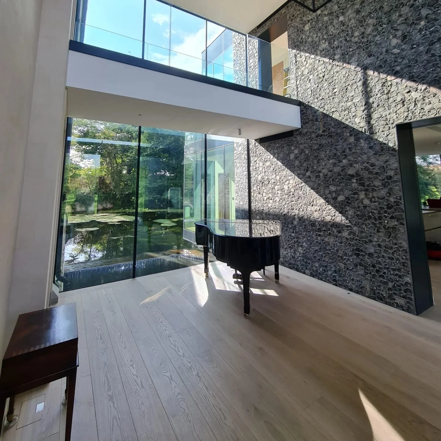 Interior of a modern house featuring a black grand piano near large glass windows with a view of a small pond and trees outside, a textured stone wall on the right, and light wooden floor.