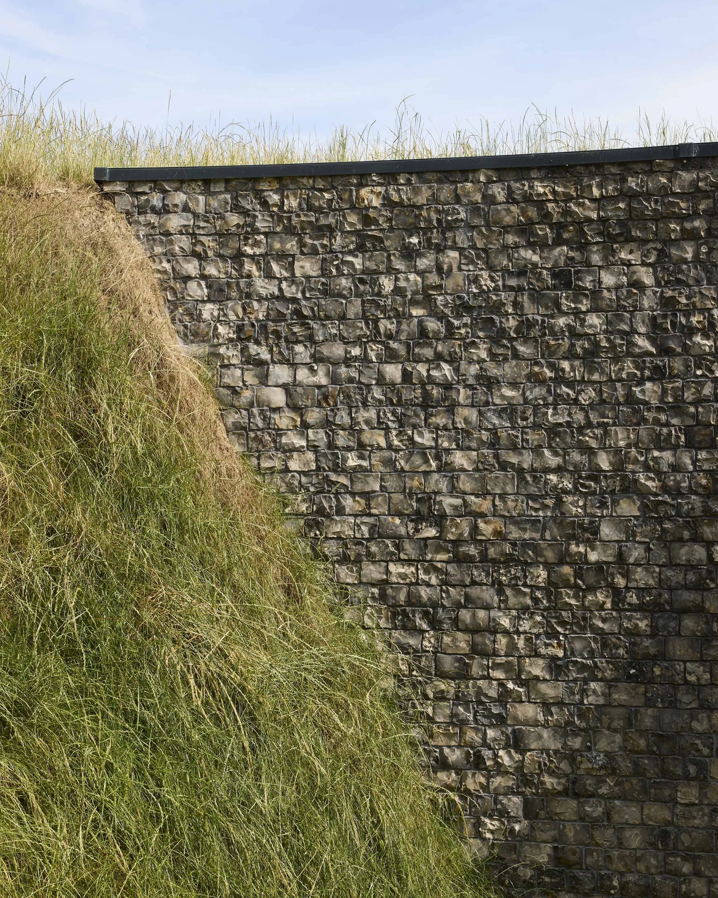 A stone and concrete retaining wall with grass and soil topped with grass growing on the earth behind it, under a blue sky.