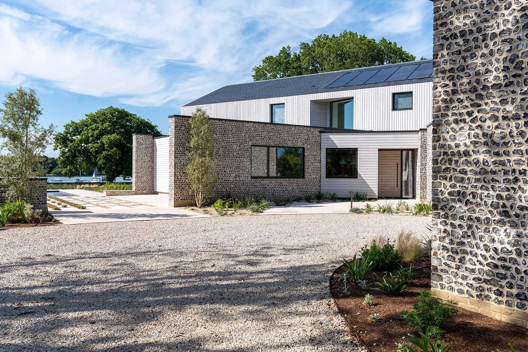 Modern house with stone and white exterior, large windows, and solar panels on roof, surrounded by landscaped yard with small plants and trees, under a blue sky with clouds.