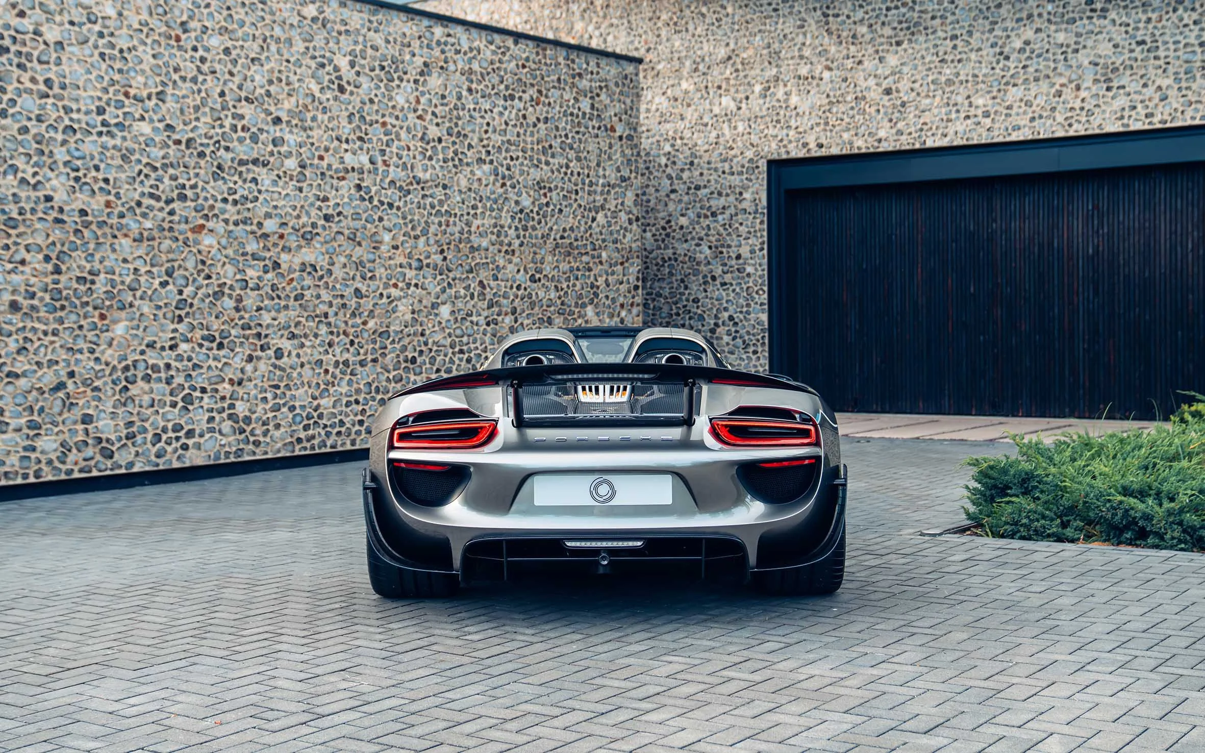 A silver Porsche sports car parked in front of a building with a pebble stone wall and a large dark garage door.