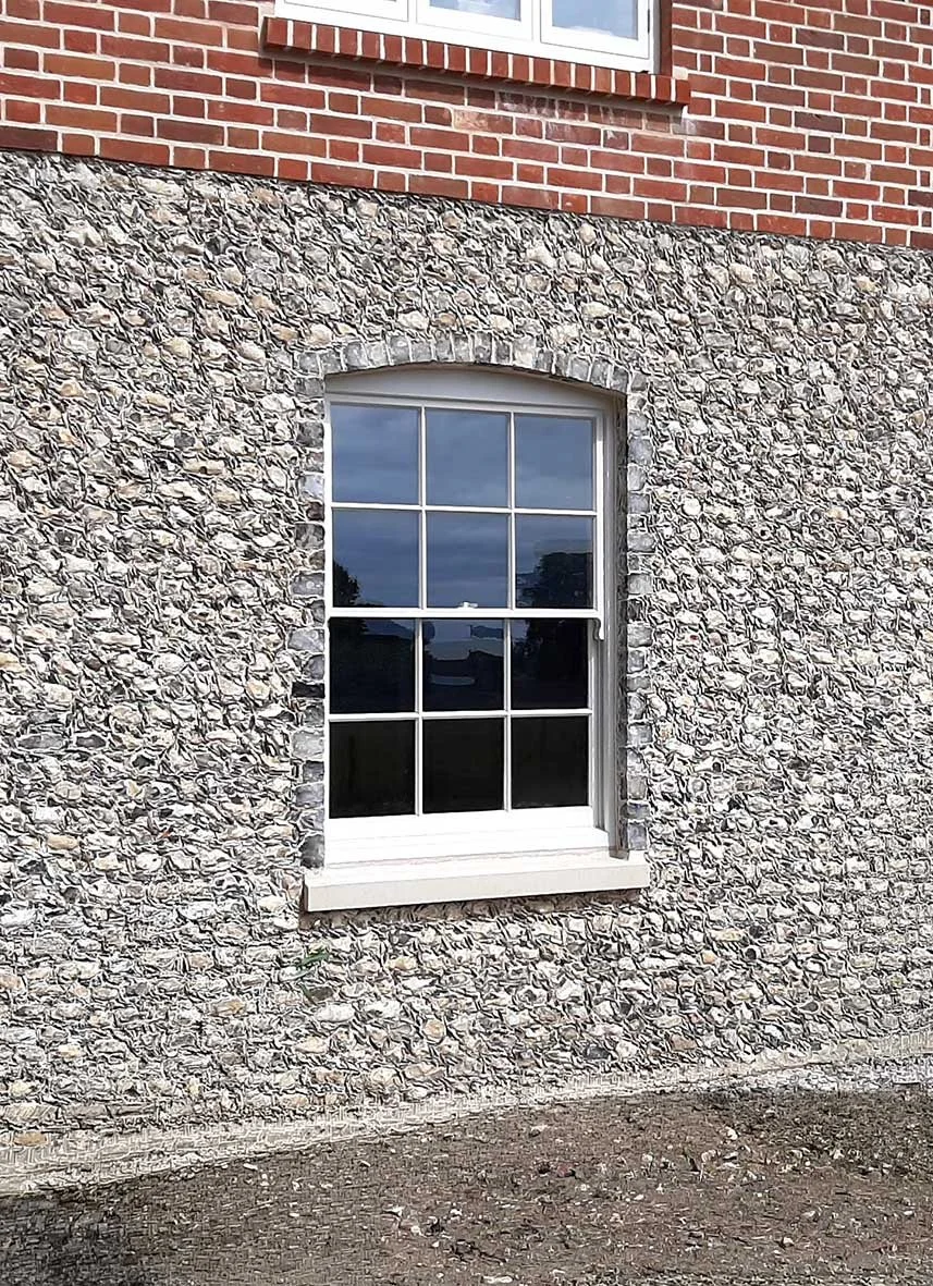 Exterior view of a brick and stone house wall with a window reflecting a cloudy sky. The window has white framing and a decorative arched brick header.