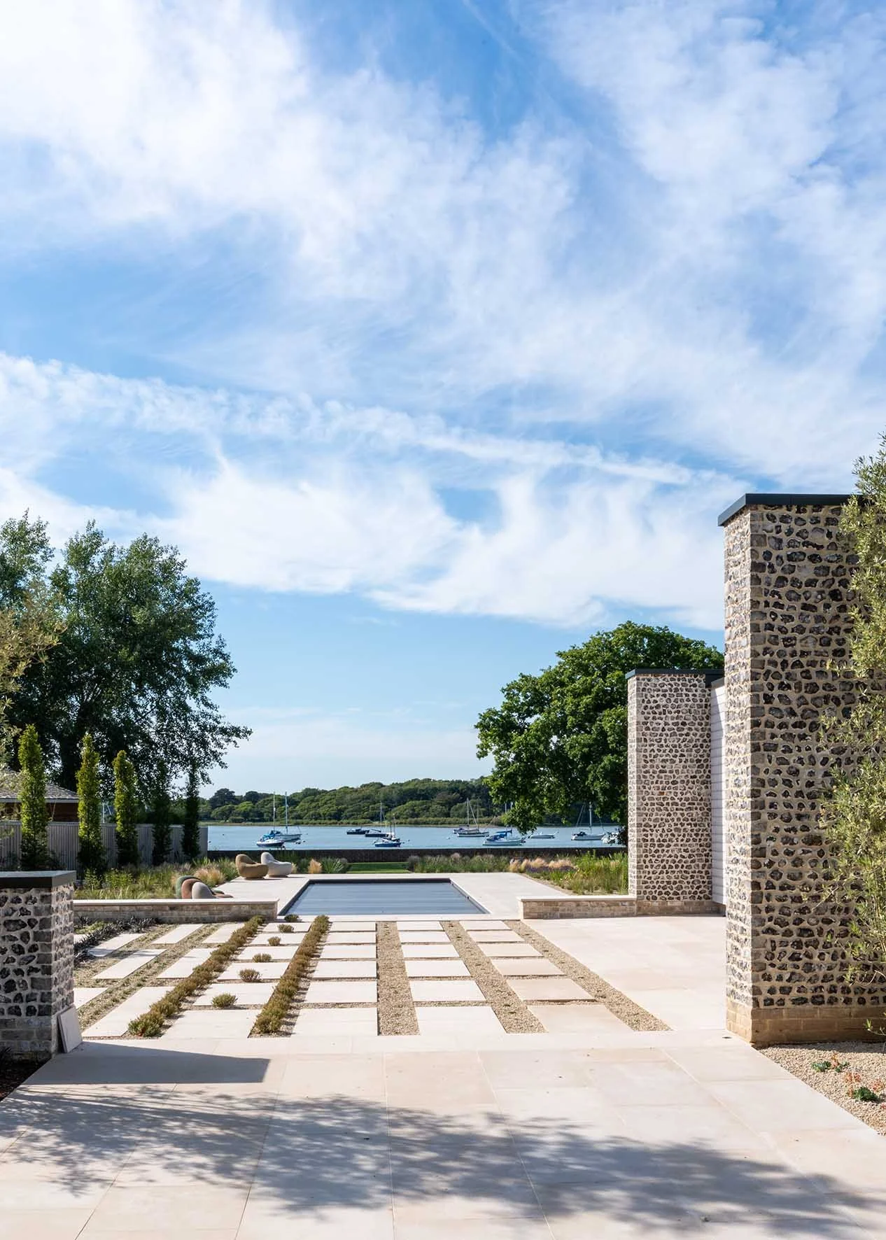 Modern outdoor patio with stone pillars, view of a lake with sailboats, and a landscaped garden under a blue sky with clouds.