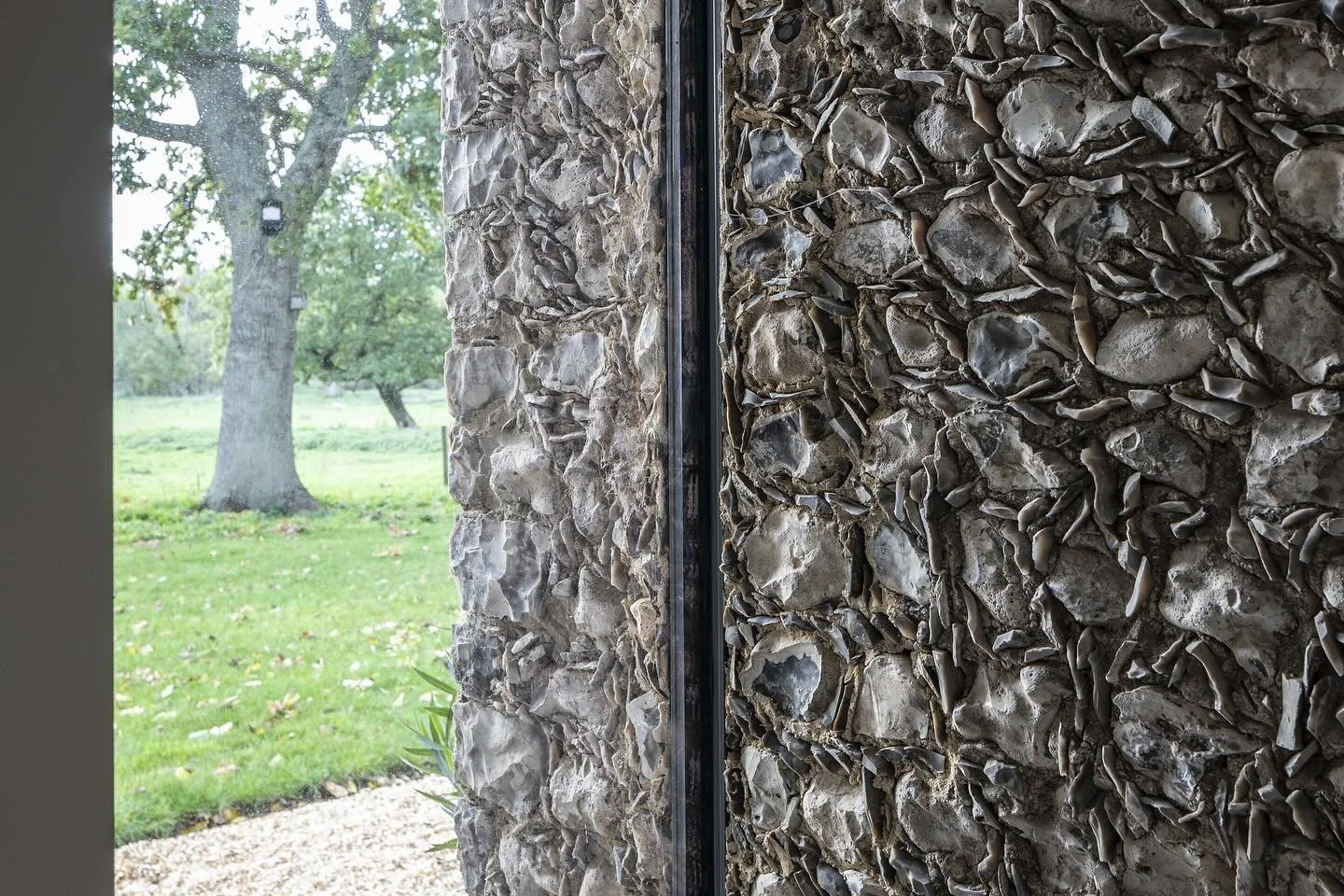 Close-up of a textured stone wall with an outdoor view of a grassy yard and trees through a glass window.