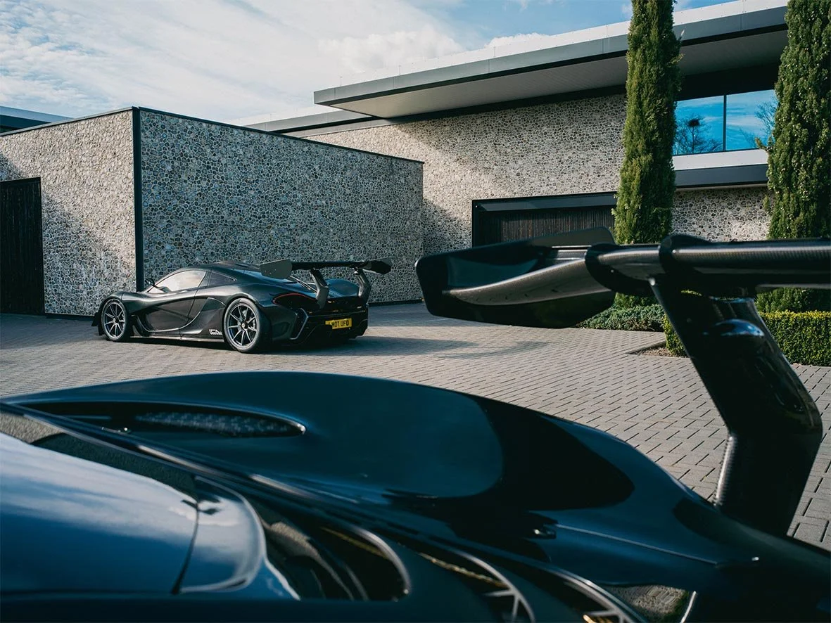 Luxury modern house with two sports cars parked outside, one with a large rear wing and the other partially visible in the foreground.