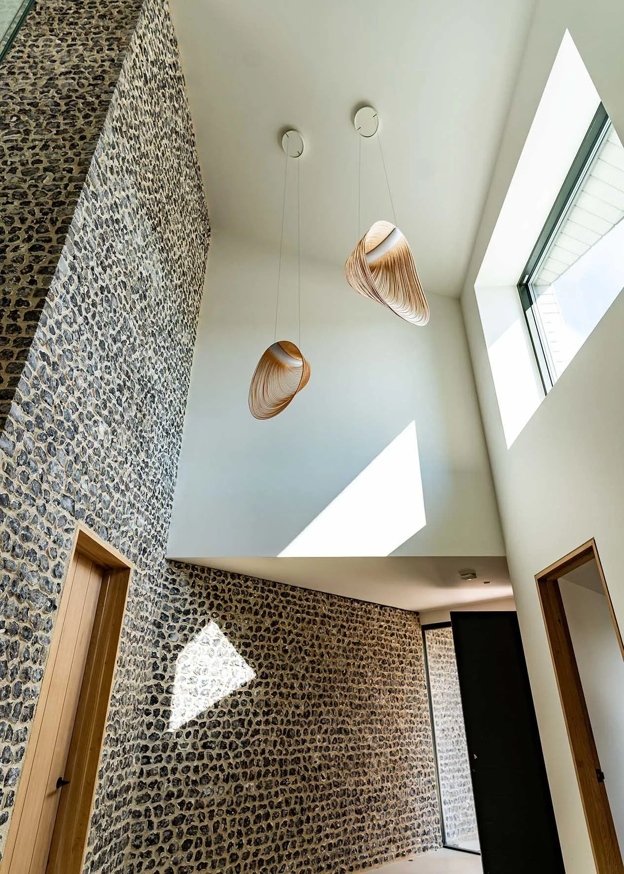 Interior view of a modern home with a high ceiling, stone accent wall, two wooden pendant lights, and a skylight allowing sunlight to enter.