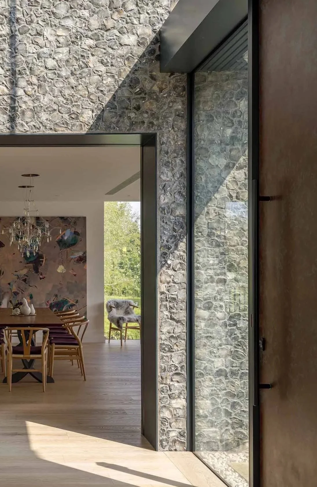 Interior view of a modern home showing a stone wall, a large glass door, and a dining area with wooden chairs and a chandelier.
