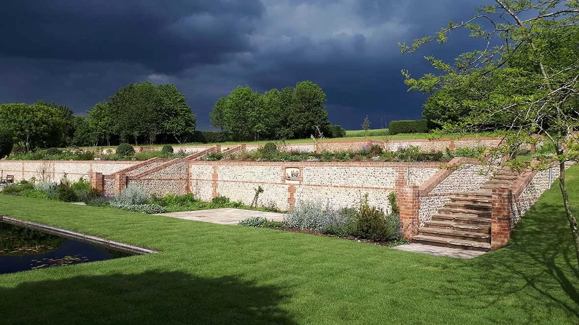 A landscaped garden with a brick and stone terraced wall, a set of brick stairs, lush green grass, trees, and a pond, under a dark cloudy sky.