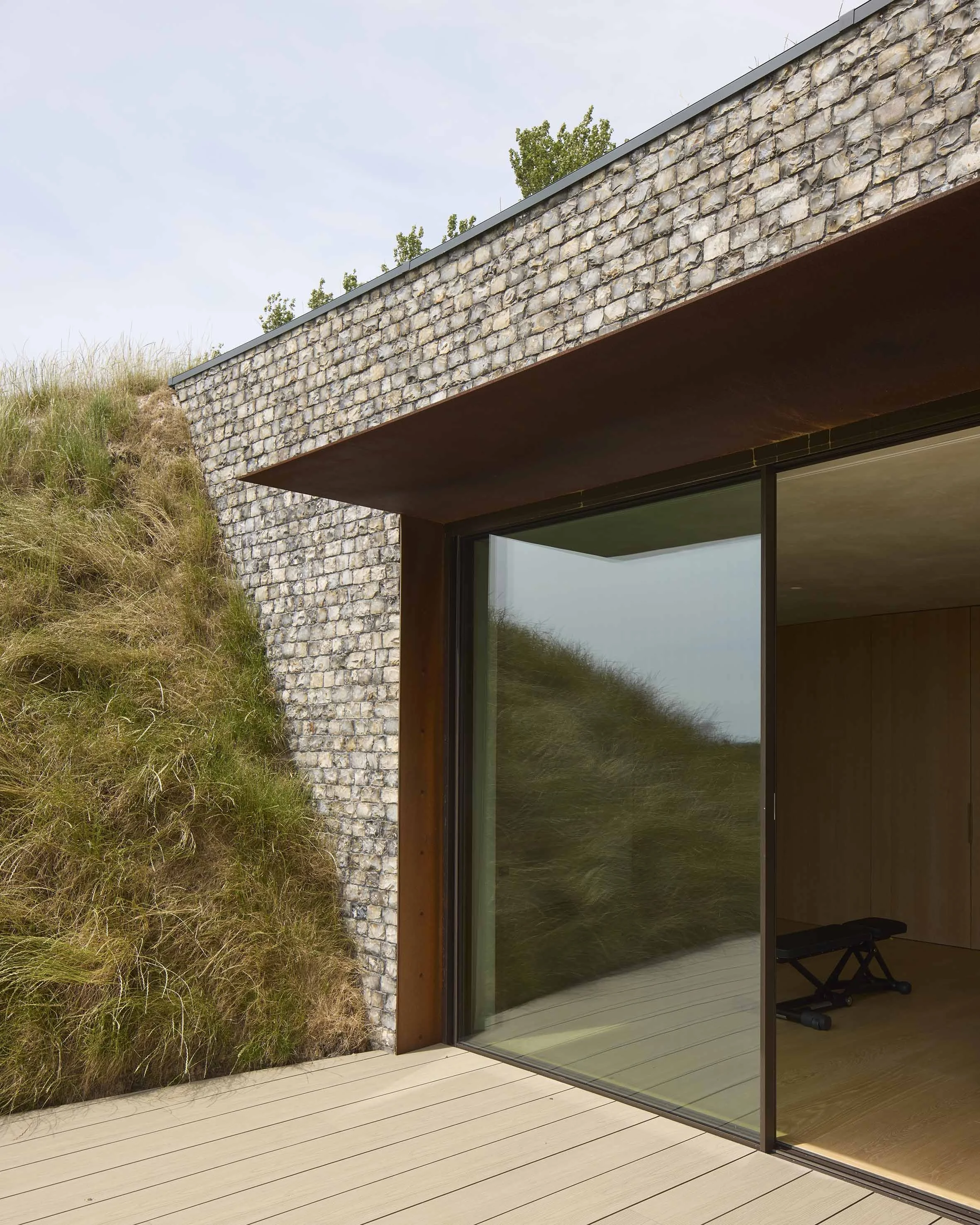 Exterior view of a modern house with a stone wall, large glass sliding door, wooden deck, and plants on a hillside.