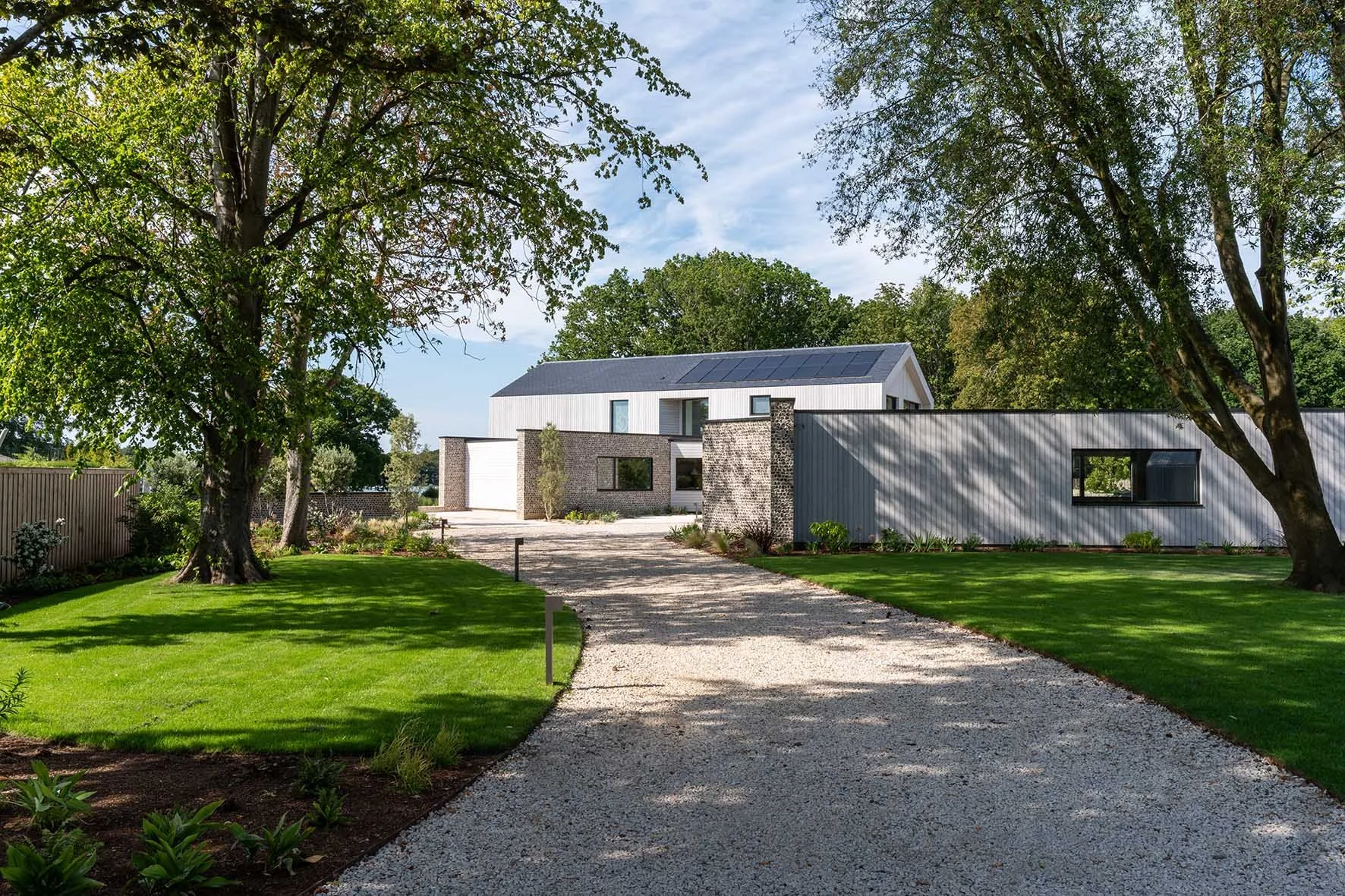 Modern house with solar panels on roof, surrounded by green trees and a well-maintained lawn, with a gravel driveway leading to the house.
