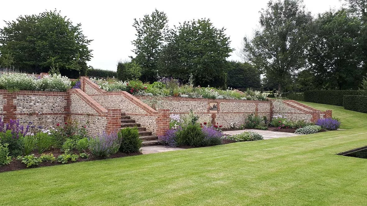 A landscaped garden with brick steps and terraces filled with colorful flowers and shrubs, set against a backdrop of large trees and a well-manicured lawn.