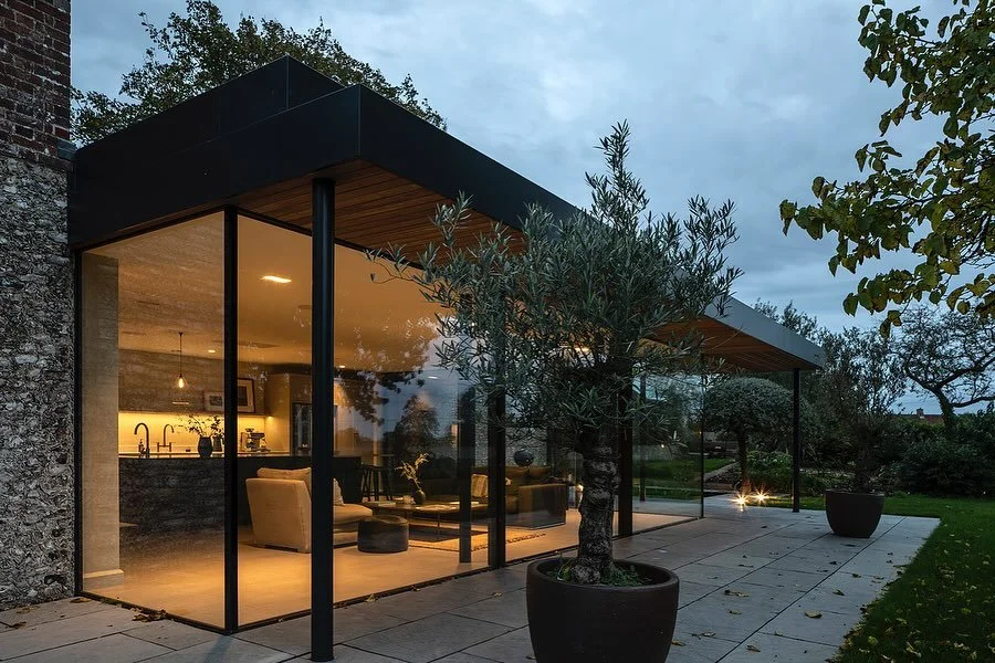 Modern glass house with an outdoor patio and potted trees at dusk