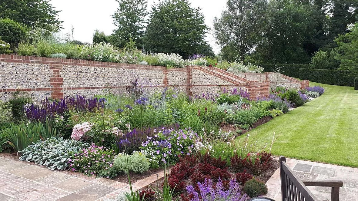 A well-maintained garden with colorful flowers, green grass, and a brick retaining wall with steps, surrounded by trees.