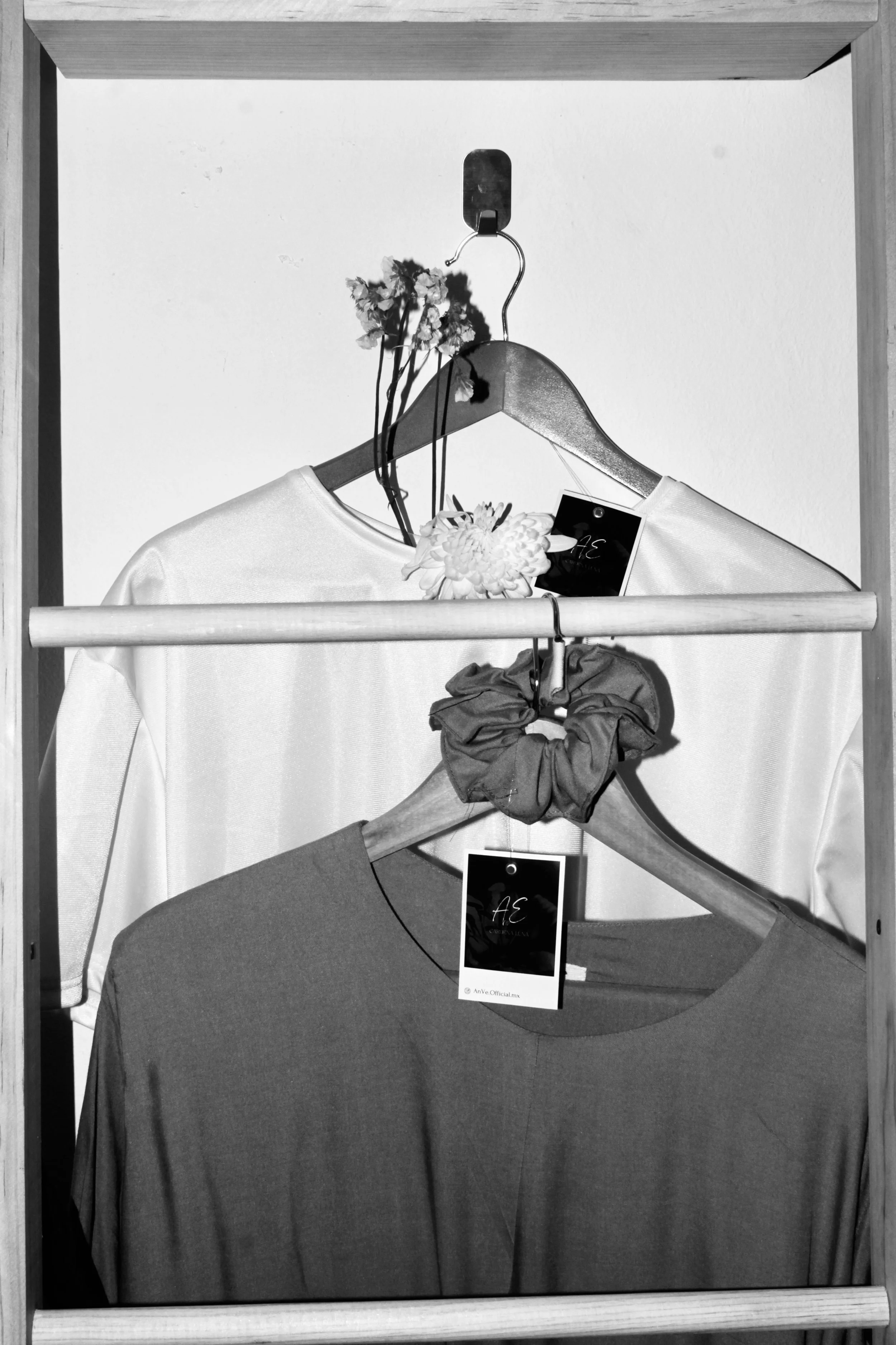 Dos camisetas colgadas en una estructura de madera, una blanca y una gris, con etiquetas y accesorios como unas flores y una cinta