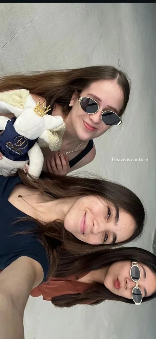 Tres mujeres sonriendo y posando para una selfie, una de ellas lleva gafas de sol y otra tiene cejas y cabello rizado, en el fondo una pared gris y en la esquina inferior derecha el texto 'Heavens couture'.