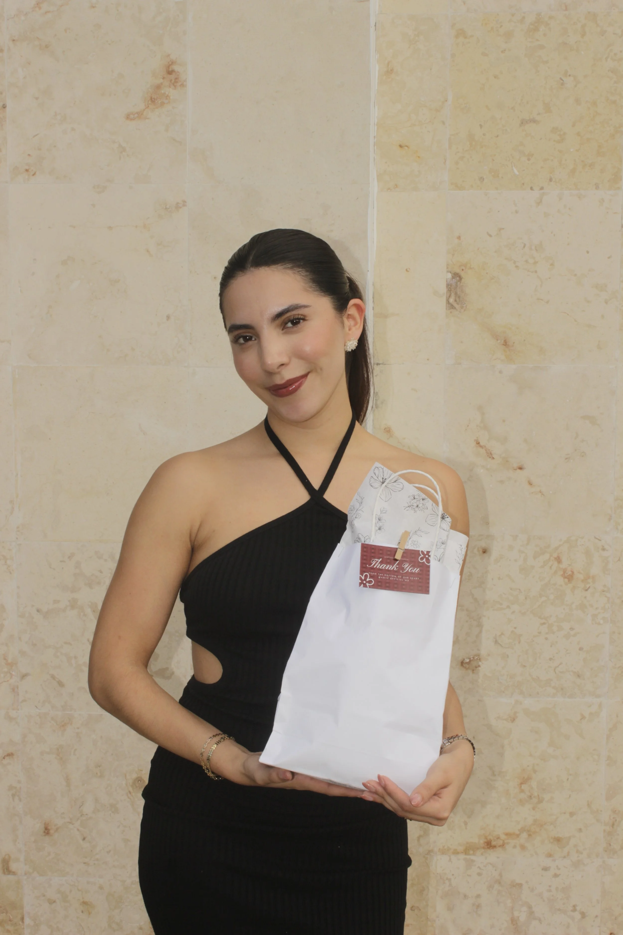 Mujer joven con vestido negro sosteniendo una bolsa blanca con una tarjeta de agradecimiento.