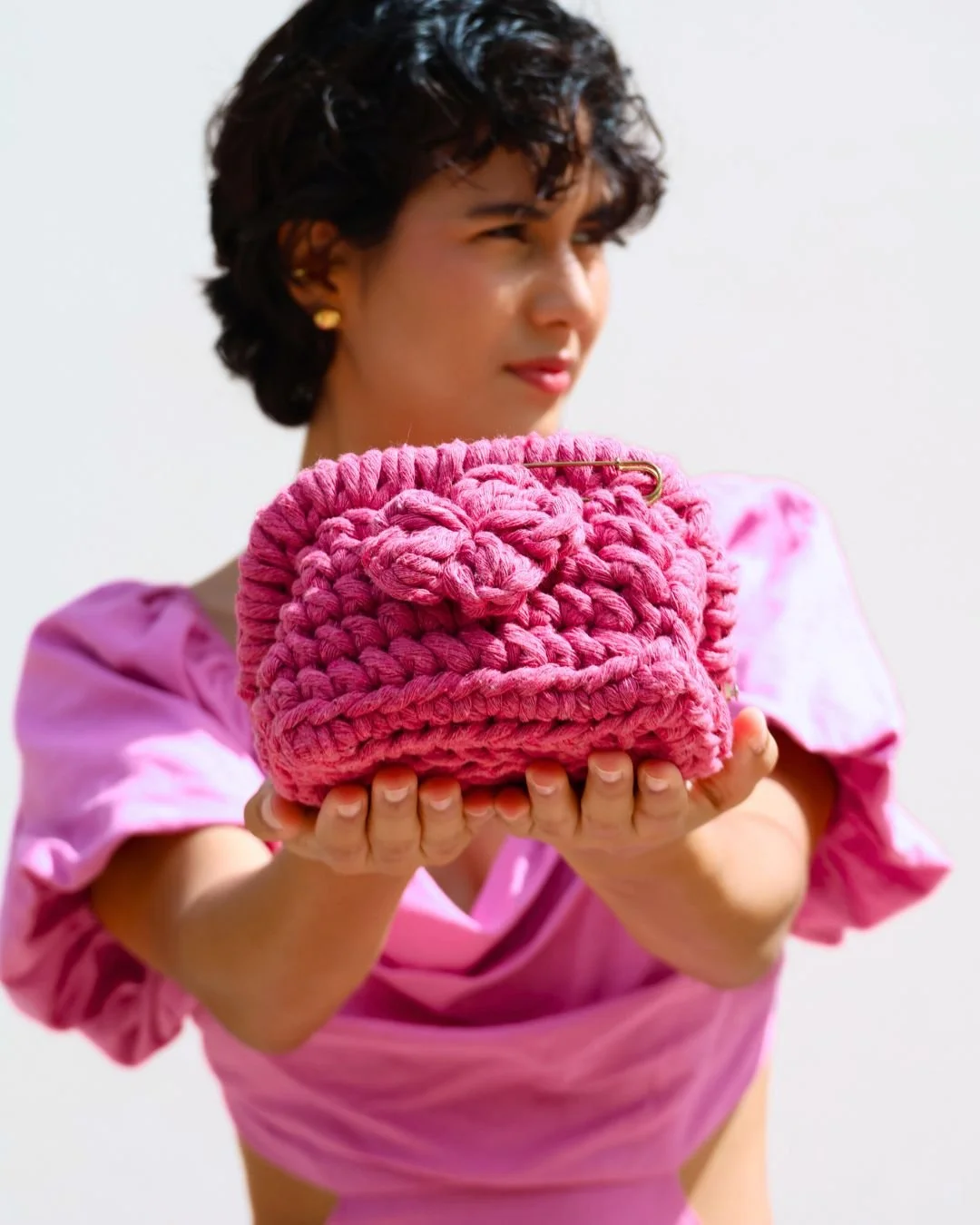 Mujer con vestido rosa sostiene una bolsa de mano rosada de crochet en forma de cara con expresión misteriosa.