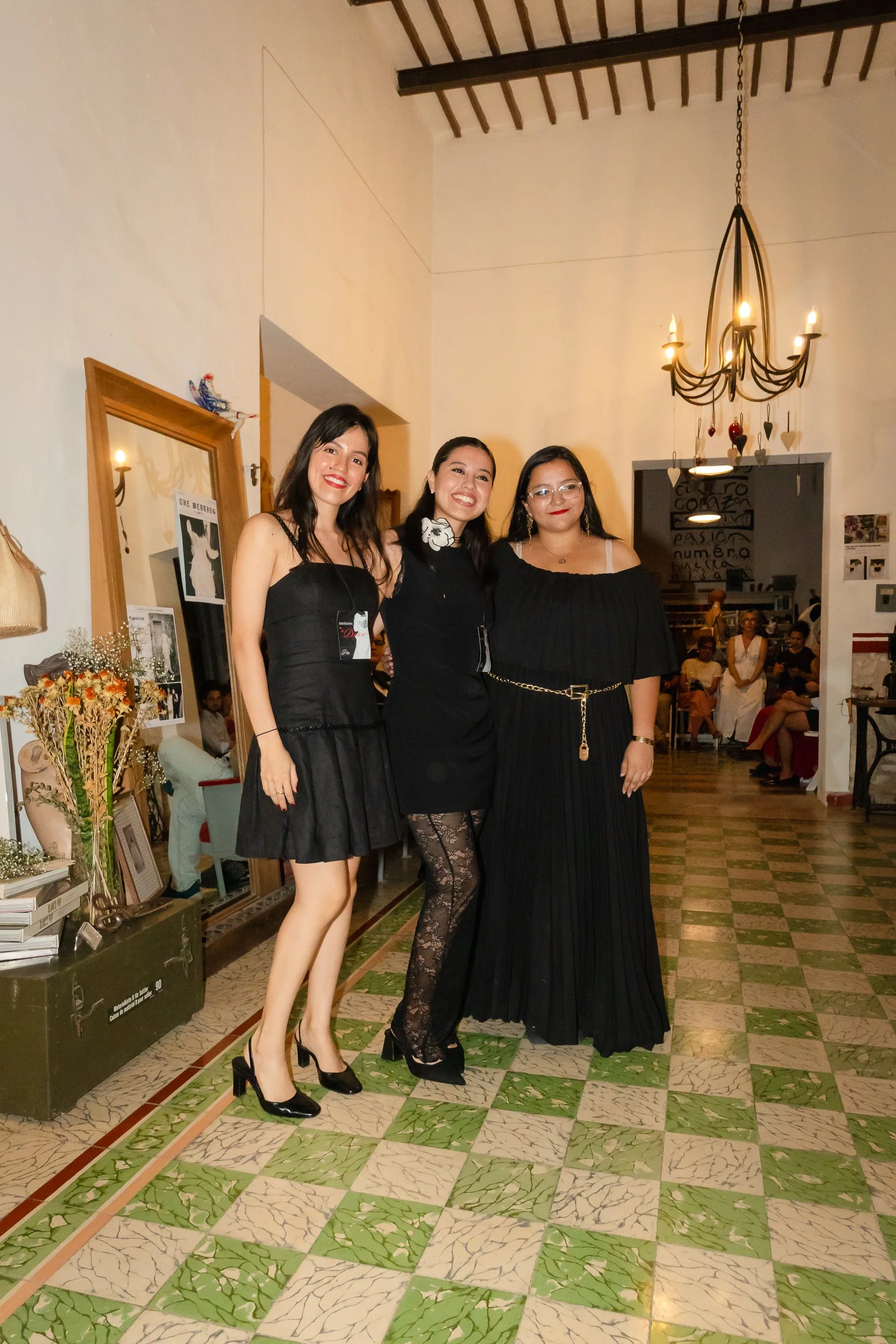 Tres mujeres vestidas con ropa negra posando en una sala decorada con flores, espejos y un candelabro, en un evento social.