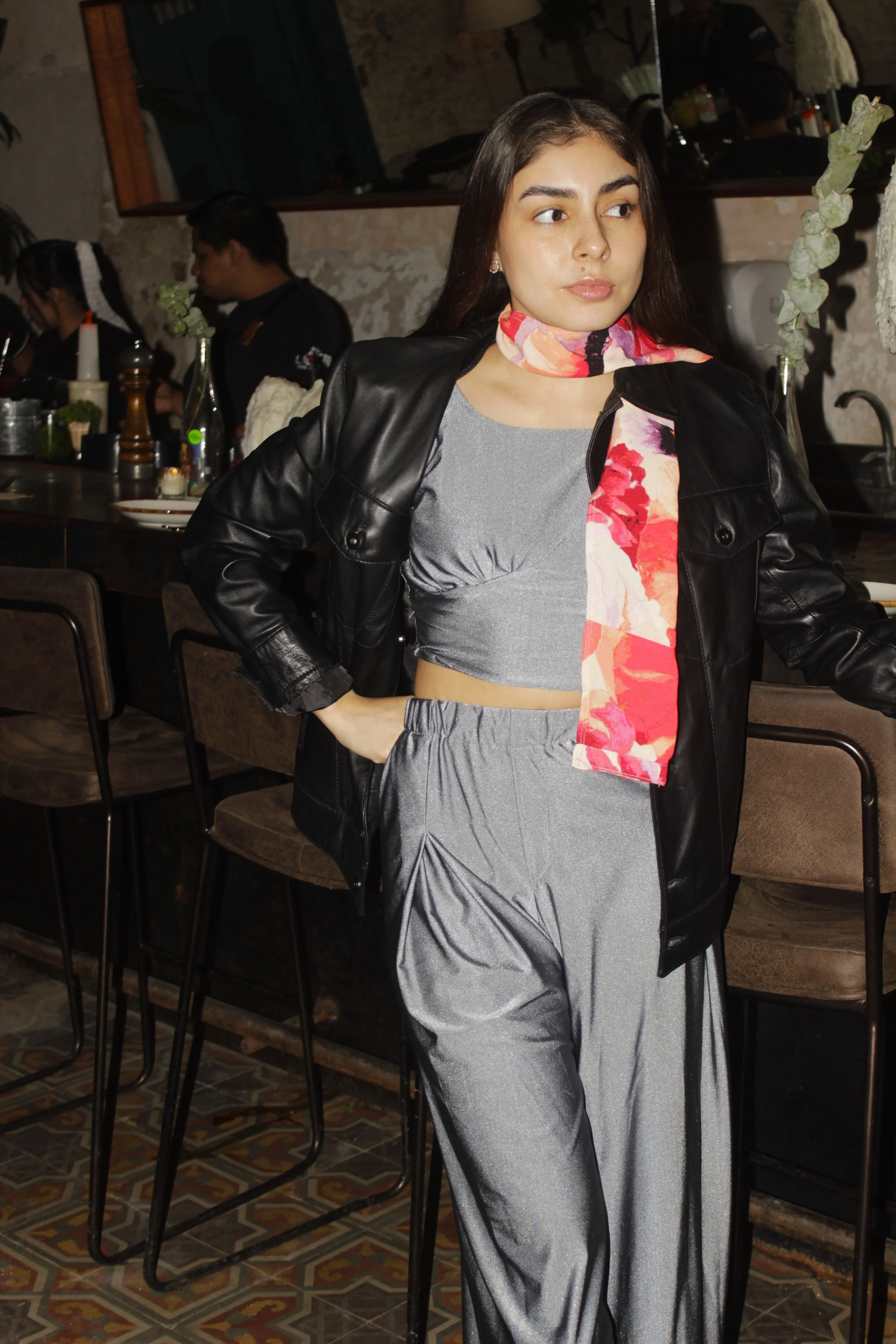 Una joven mujer con cabello largo y piel clara, viste un conjunto de pantalón y top gris, una chamarra de cuero negra, y un pañuelo colorido en el cuello, posando en un restaurante con mesas y otros clientes de fondo.
