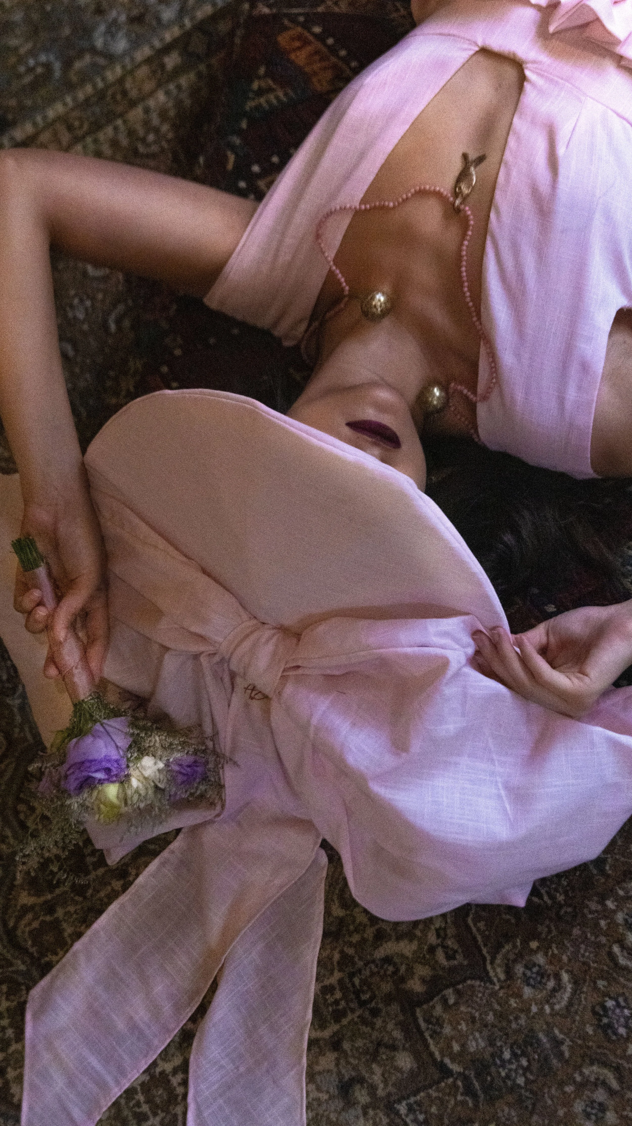 Mujer desnuda con vestido rosa y sombrero, acostada en alfombra, con collar de perlas y holding flores.