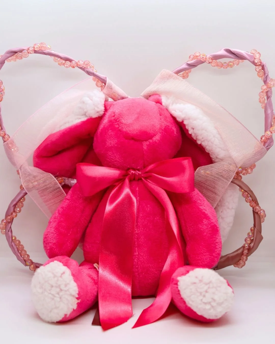 Un conejito de peluche rosa con lazos, rodeado por unas alas de mariposa decorada con cinta y perlas, en fondo blanco.
