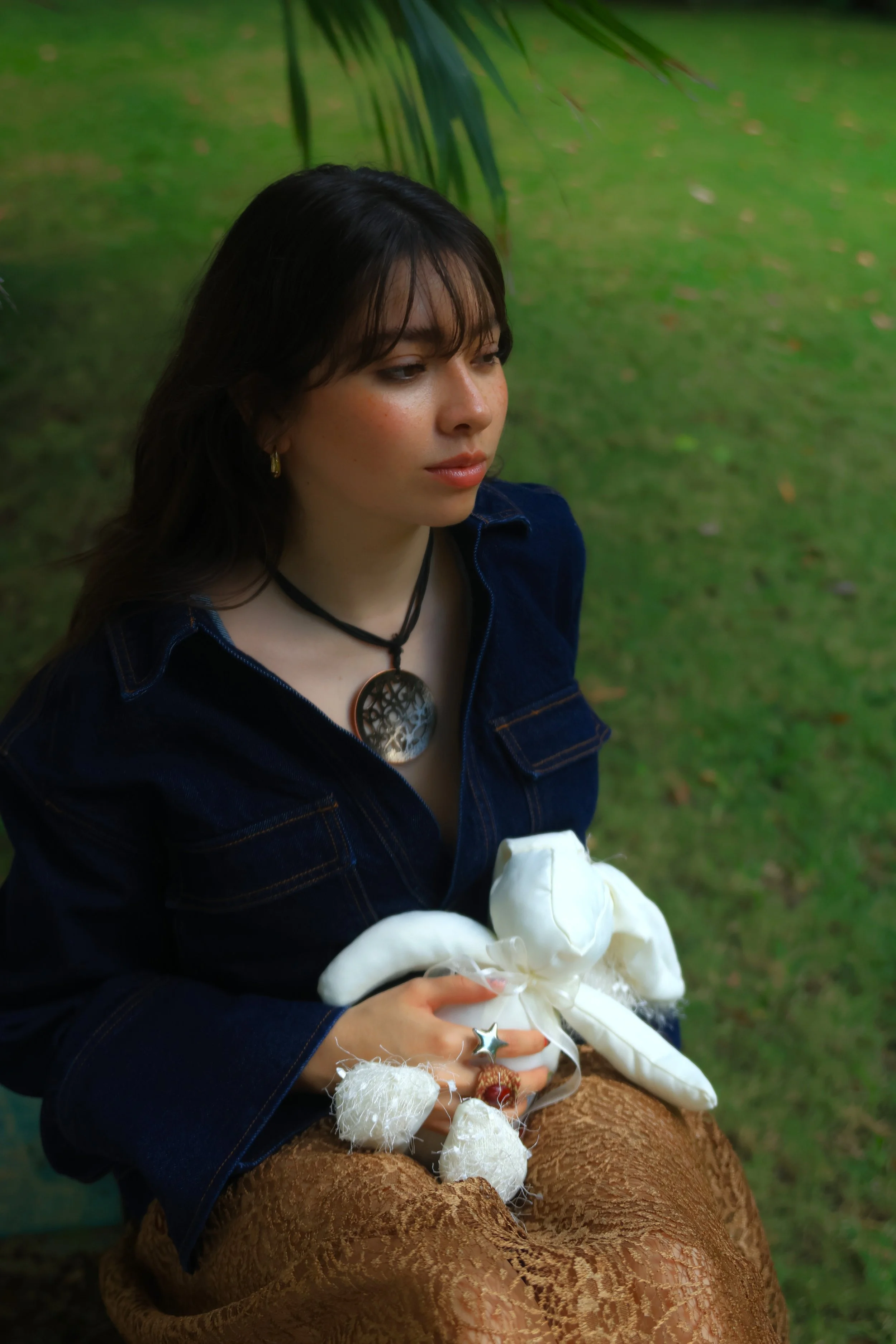 Mujer con cabello oscuro y cabello de flequillo, usando collar con colgante de árbol, vestido de mezclilla y falda marrón con patrón. Está sentada en un entorno natural con césped y plantas. Ella sostiene objetos decorativos, incluido un conejito de 