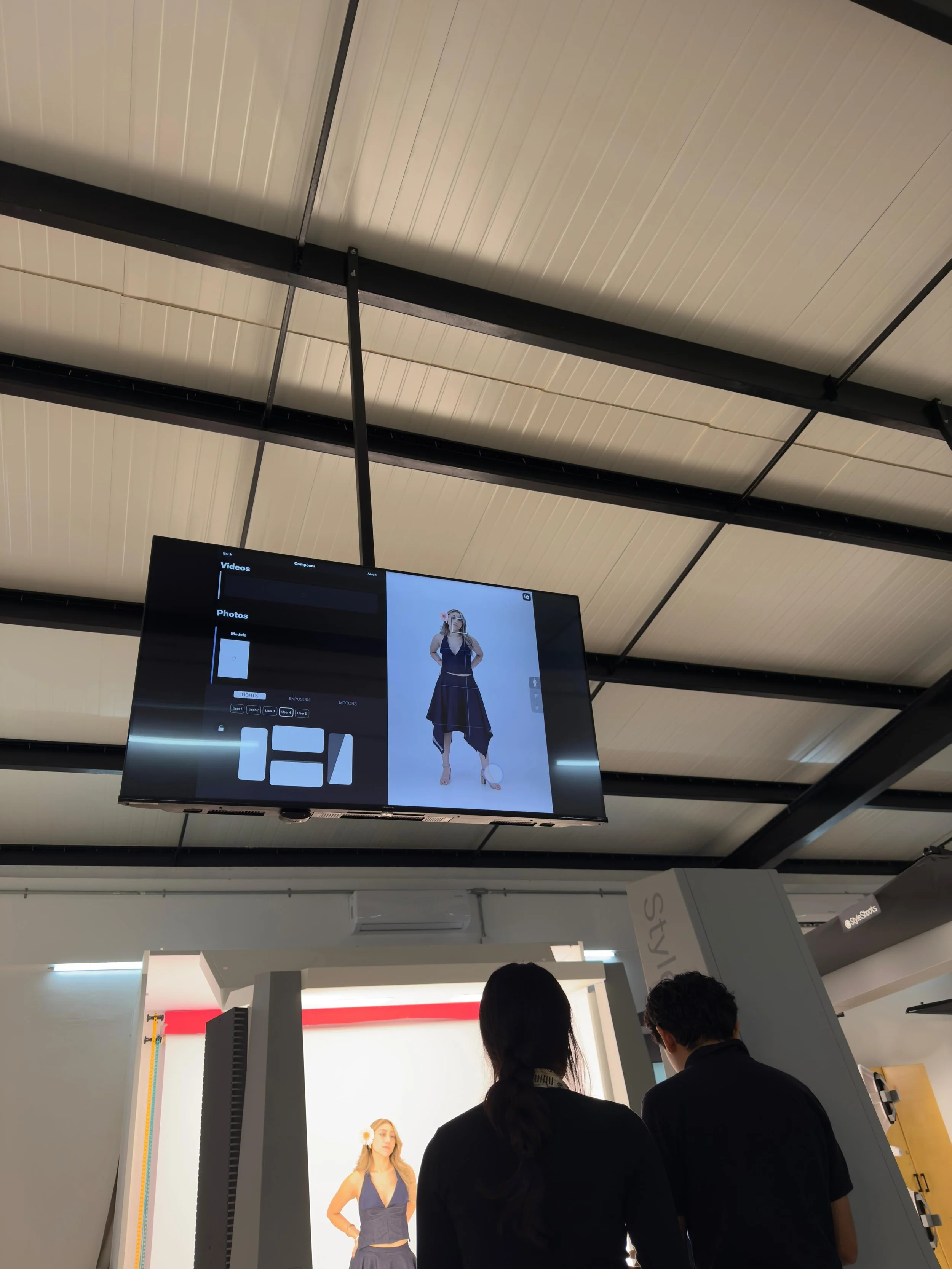 Dos personas observan una pantalla que muestra una foto de una mujer con un vestido largo y oscuro, en un entorno que parece una tienda o sala de exhibiciones de moda.