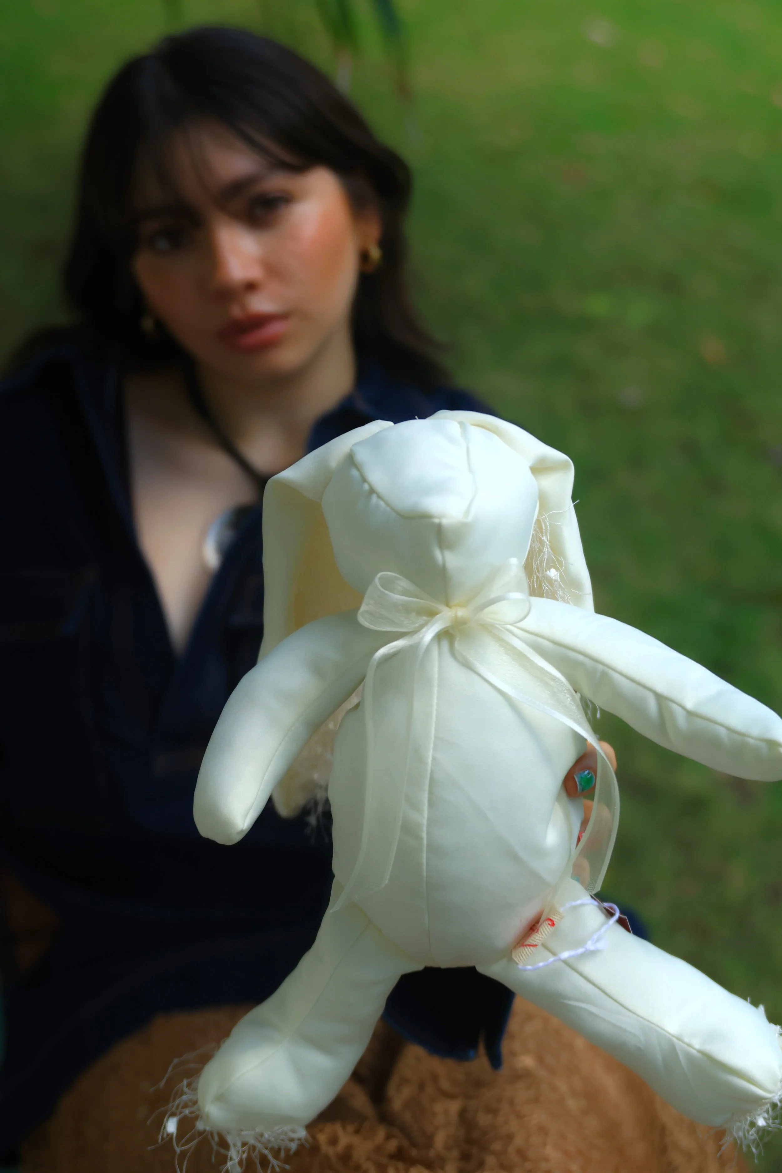Mujer con cabello largo y oscuro sosteniendo un oso de peluche blanco con lazos de tela en un fondo verde borroso.