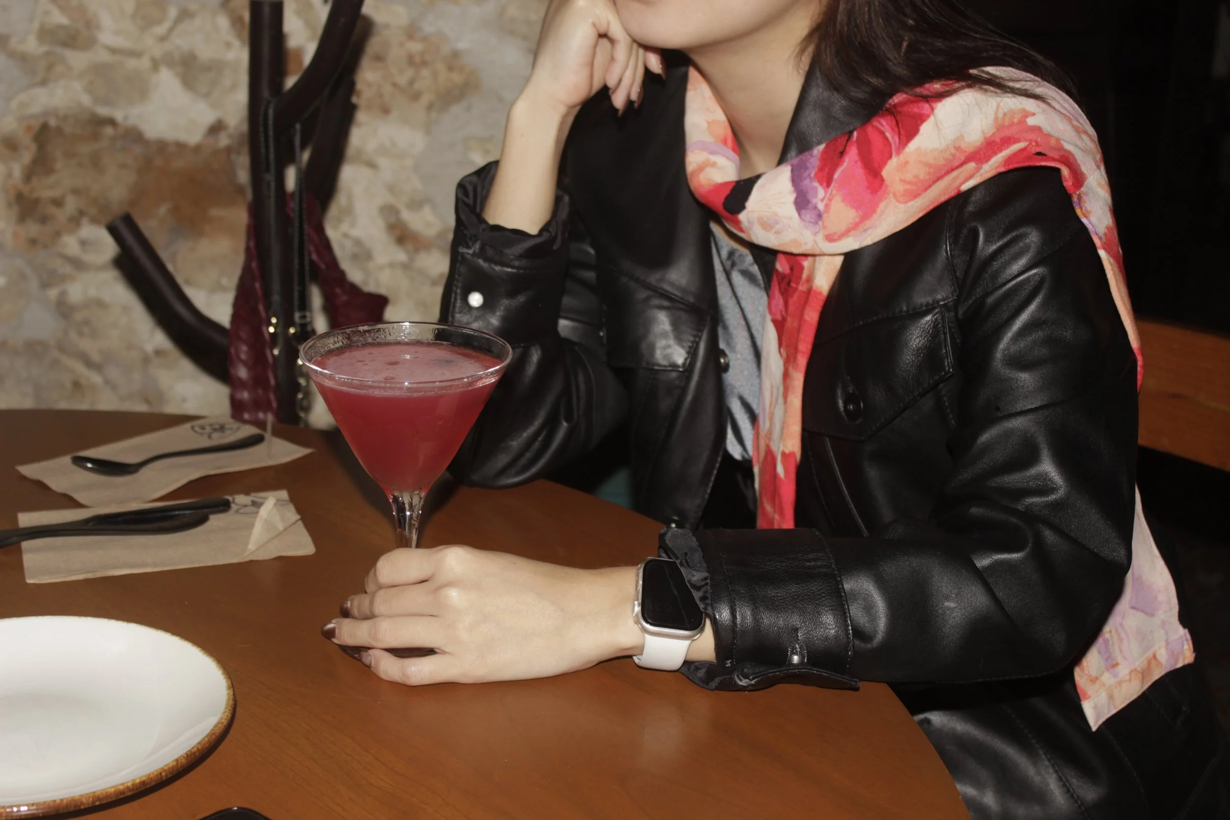 Mujer con chaqueta de cuero negro y bufanda color coral sentado en una mesa de restaurante, sosteniendo una copa de cóctel rosado, con accesorios como reloj inteligente, en un ambiente con pared de piedra.