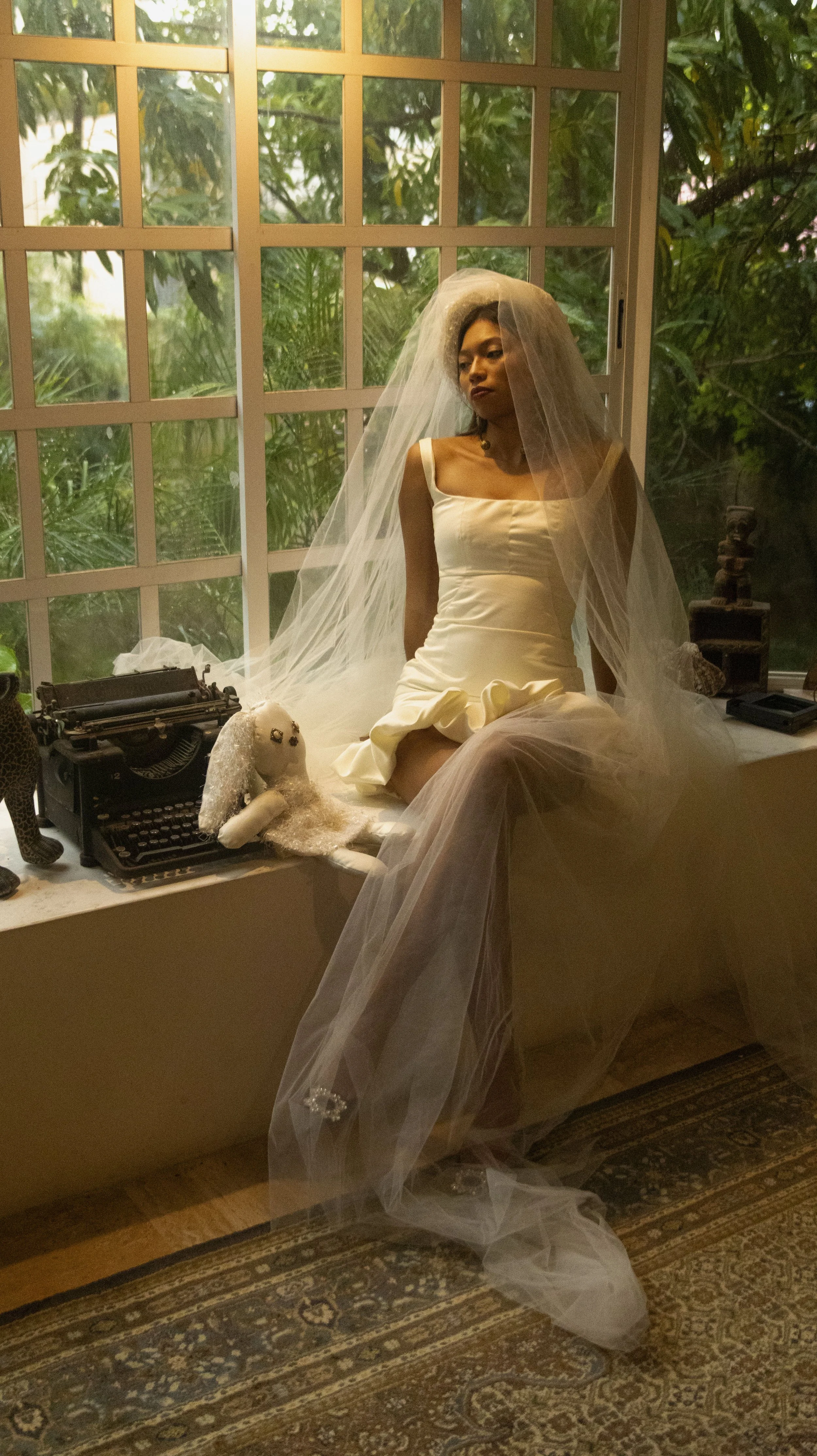 Mujer en vestido de novia con velo sentado en una repisa junto a una máquina de escribir y muñeco de peluche en una habitación con ventanas grandes y persianas.