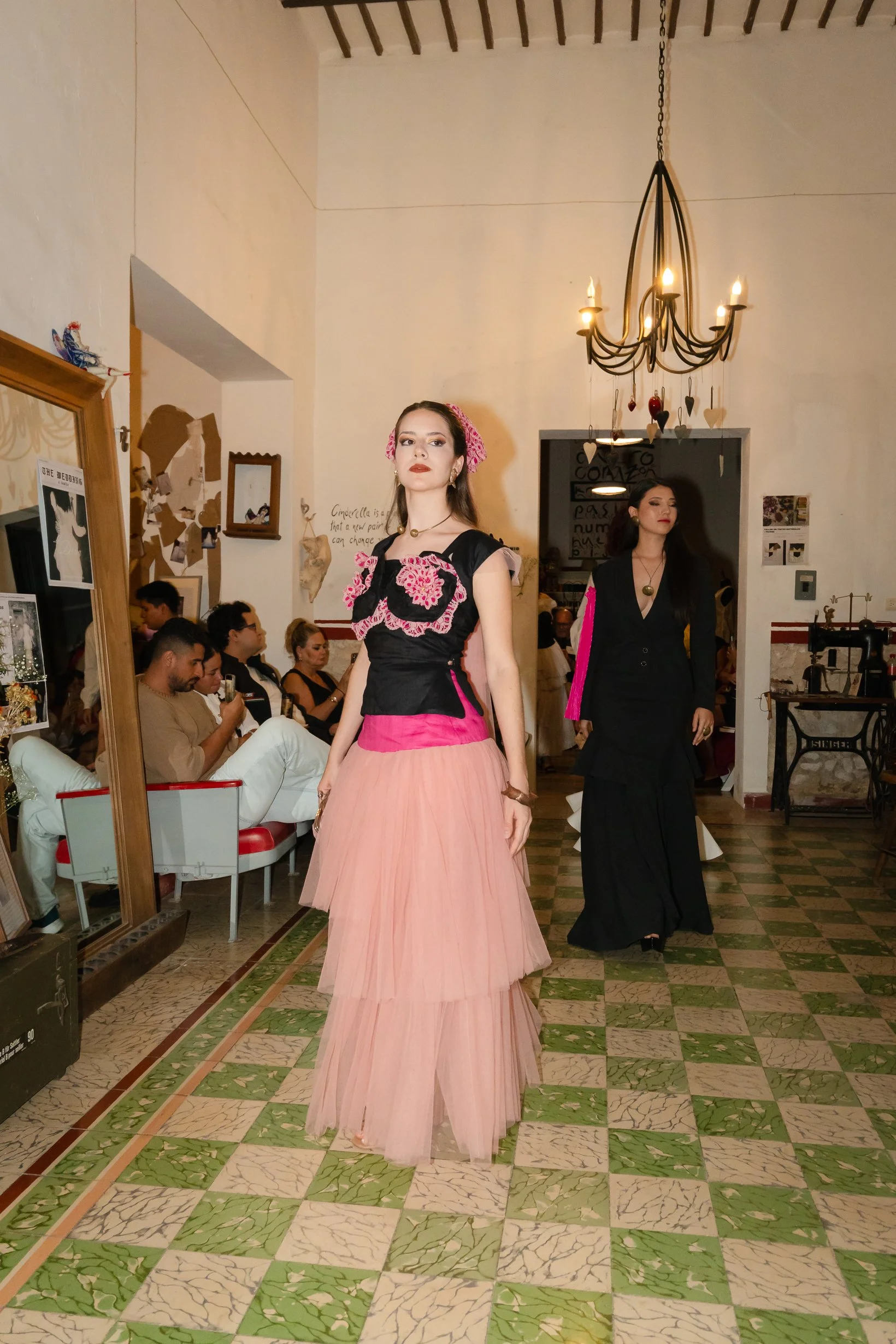 Modelo desfilando en una pasarela con un vestido largo rosa y detalles en negro y rosa, en un ambiente de salón con chimenea y personas sentadas observando, decoraciones colgantes y lámpara de araña.