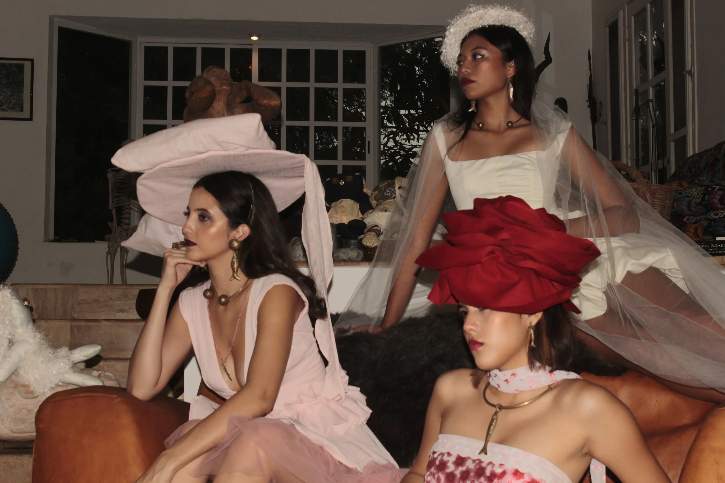 Tres mujeres vestidas con atuendos elegantes y accesorios llamativos en un entorno decorado con objetos artísticos y textiles coloridos.