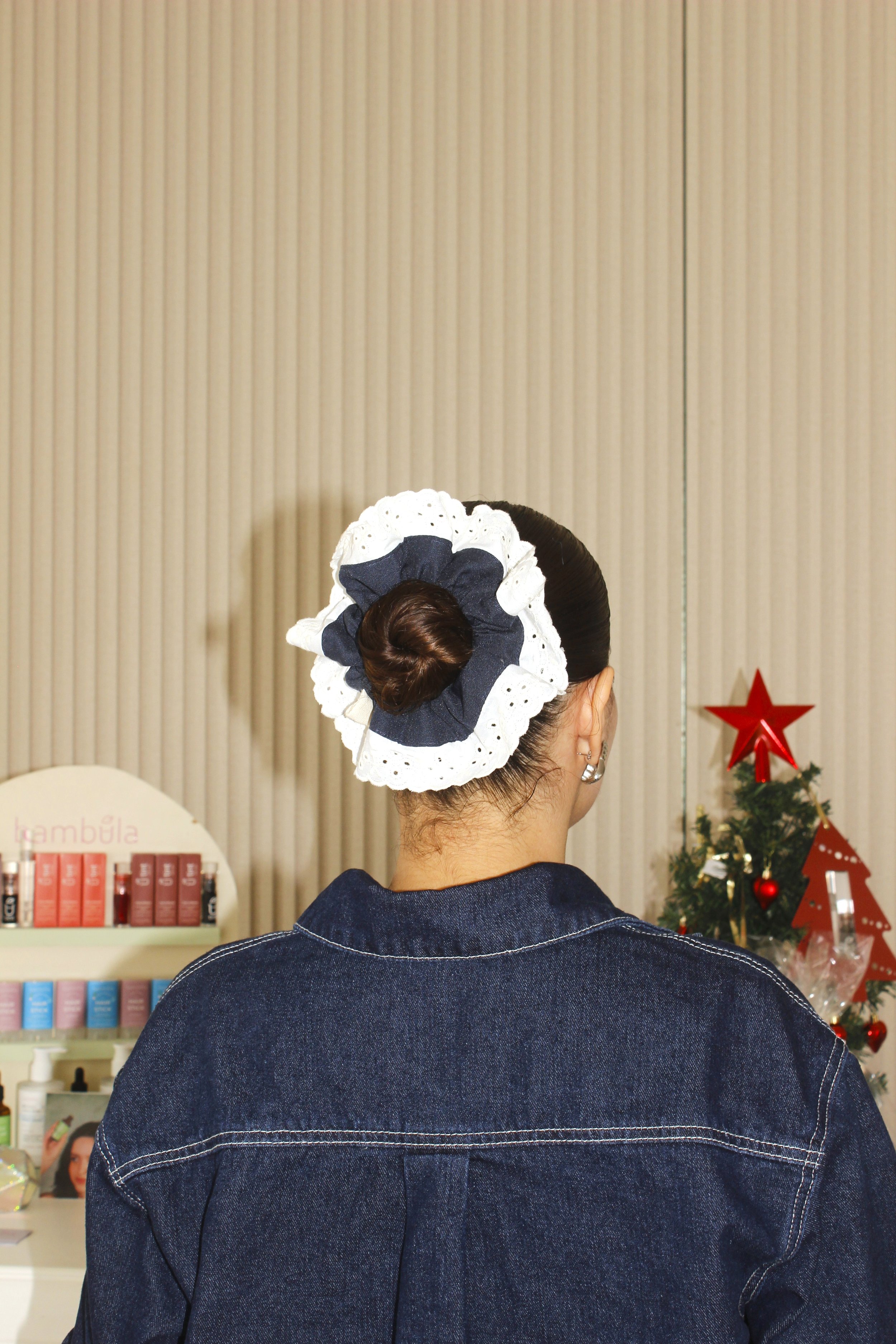 Mujer de espaldas con peinado en moño y lazos grandes, usando chaqueta de mezclilla, en un lugar con decoraciones navideñas.