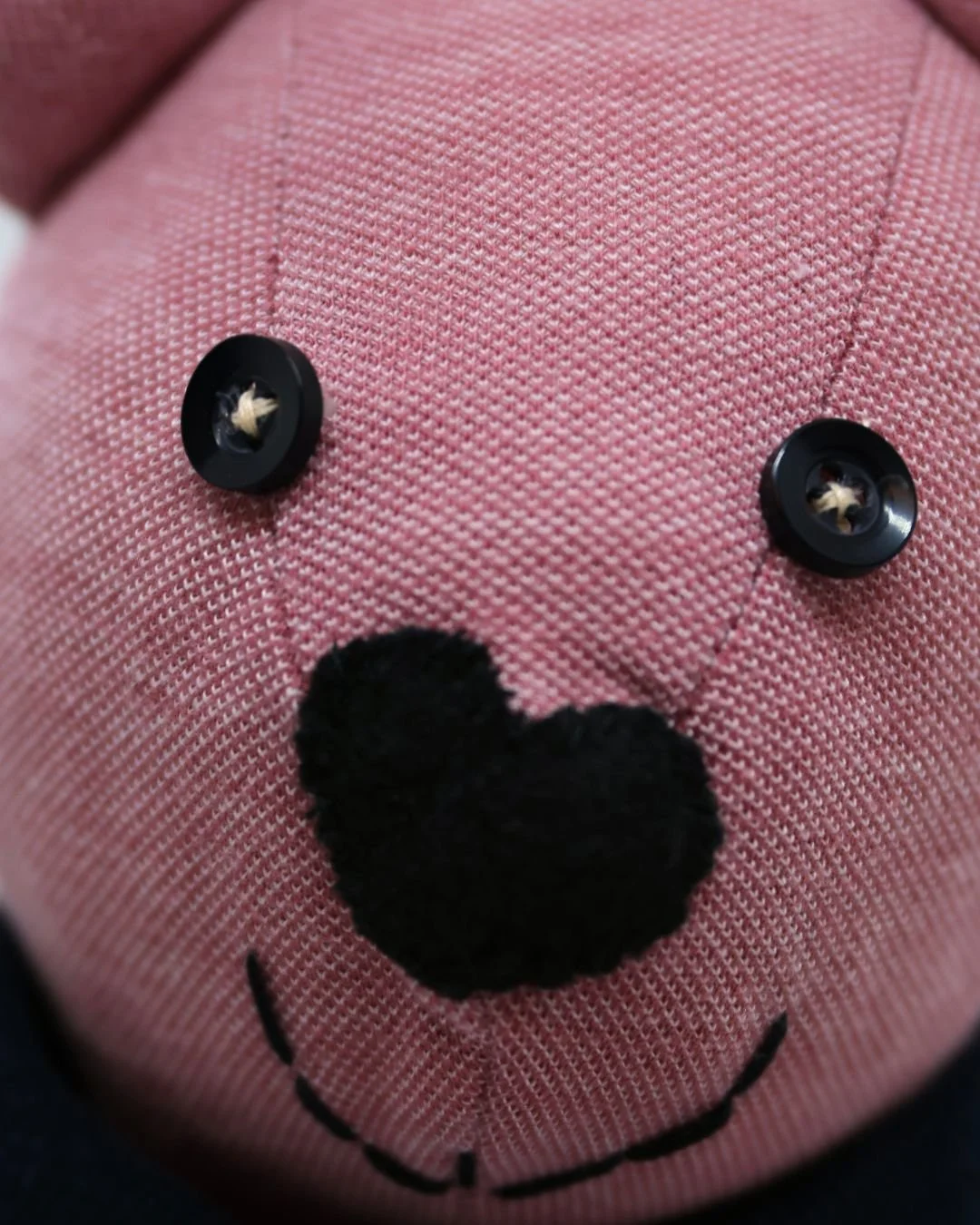Primer plano de una carita de peluche rosa con botones negros en forma de ojos, una nariz de fieltro negro y una sonrisa bordada.
