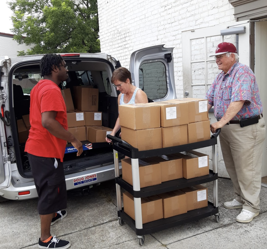three people are standing behind a van loading brown cardboard boxes inside